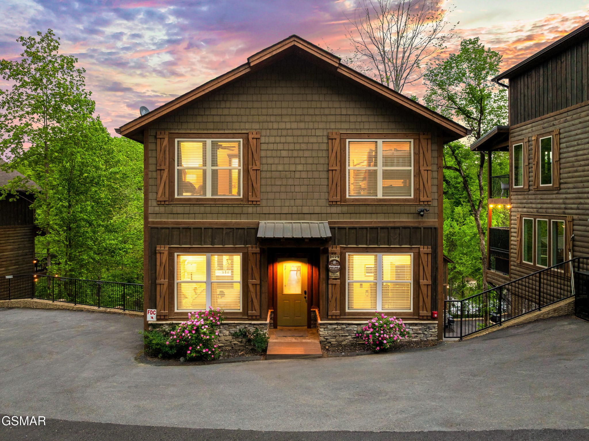 Two-story timber-style home with glowing windows, pink flowering shrubs, and a colorful dusk sky.