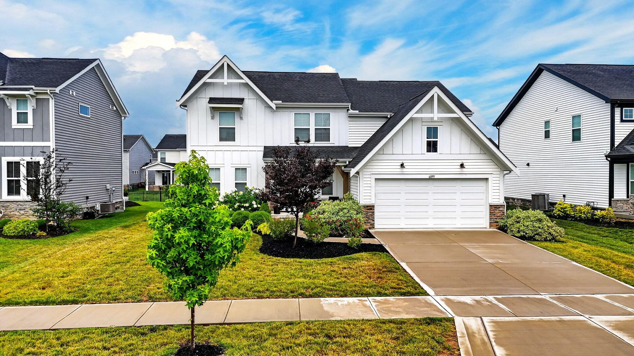 White two-story house with a two-car garage, landscaped shrubs, and a wide concrete driveway.