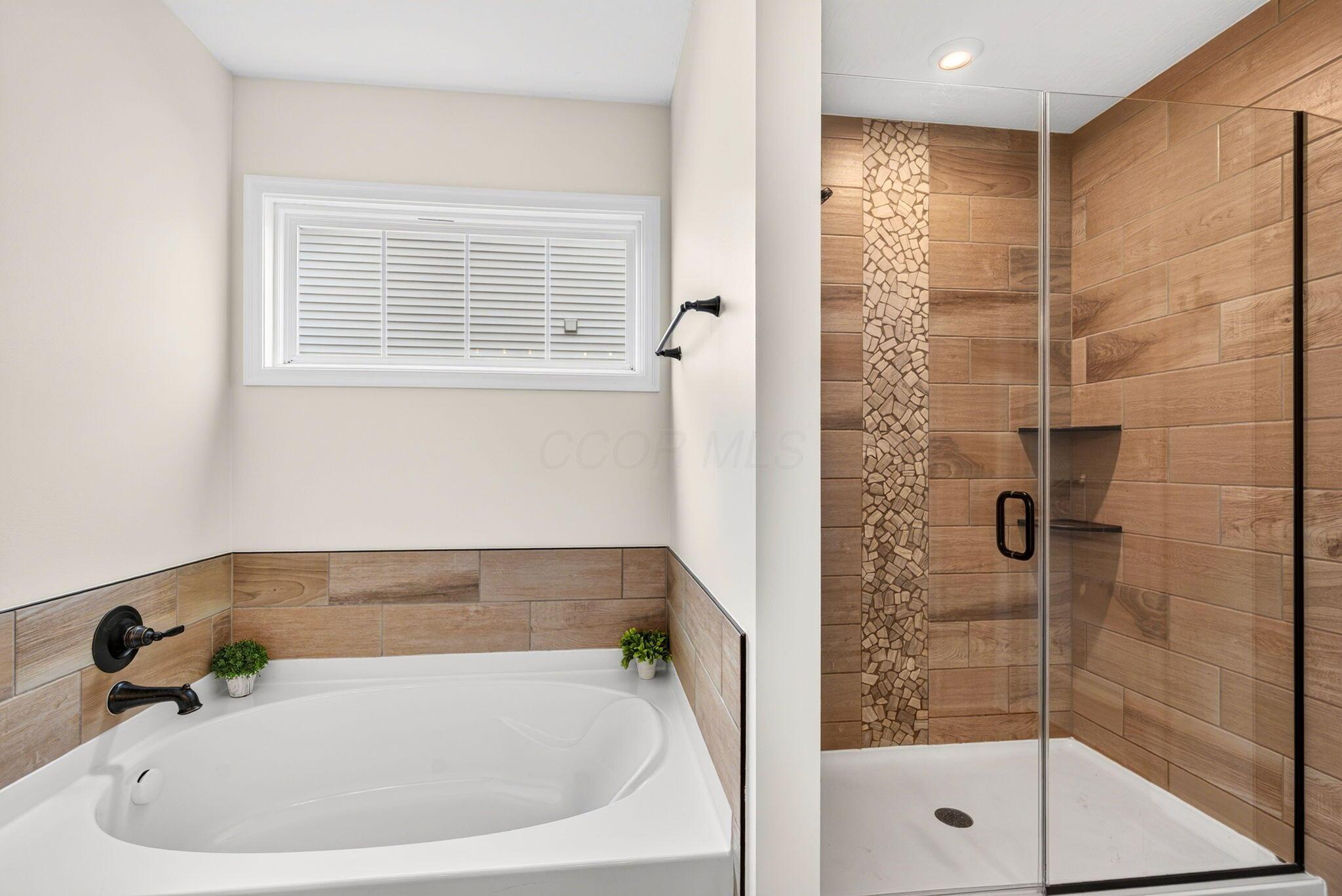 Bathroom with a soaking tub and glass shower, both finished in warm wood-look tile.