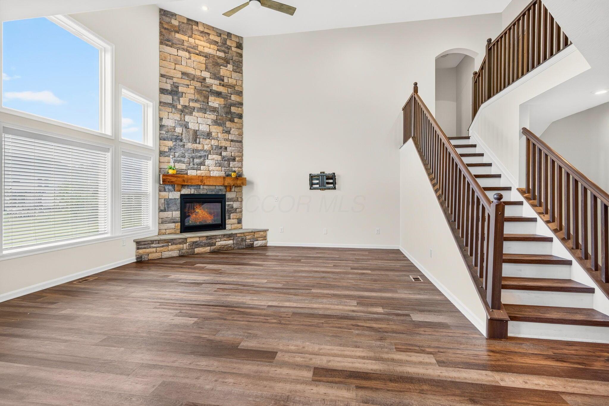 Living room with a stone fireplace, tall windows, wood-look floors, and a dark-railed staircase.