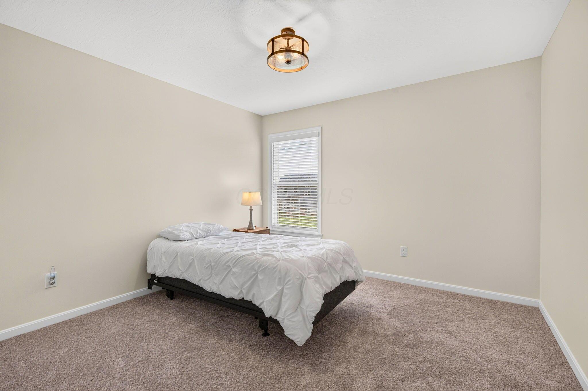 Staged bedroom with white bedding, beige carpet, and a window with white blinds.