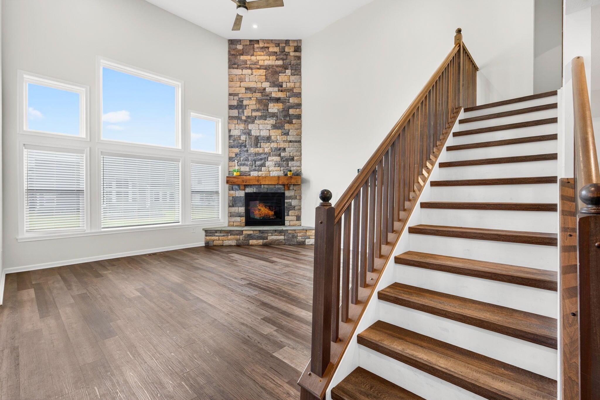 Living room with a stone fireplace, large windows, and a dark wood staircase.
