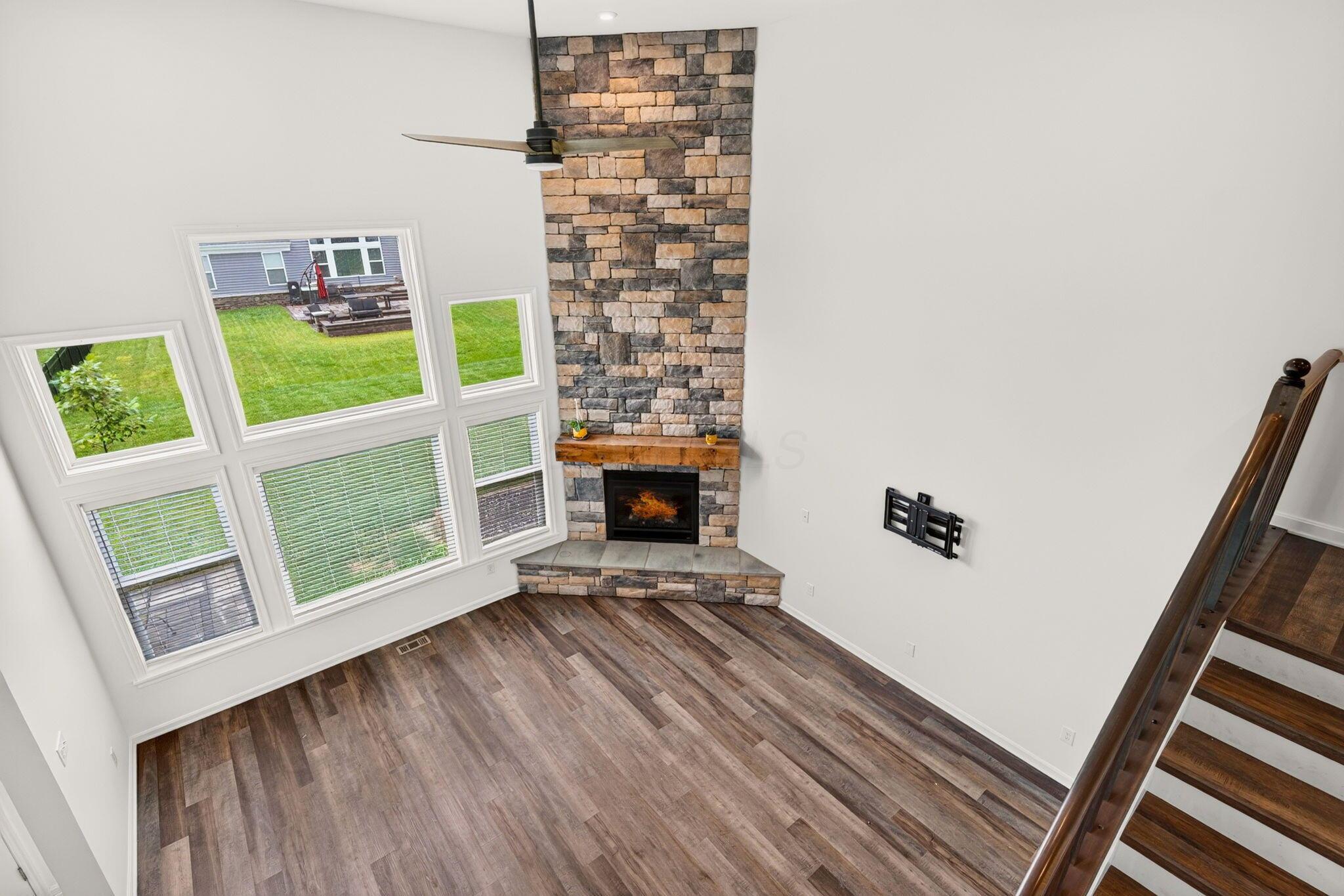 Vacant living room with a tall stone fireplace, large windows, and dark wood-look flooring.