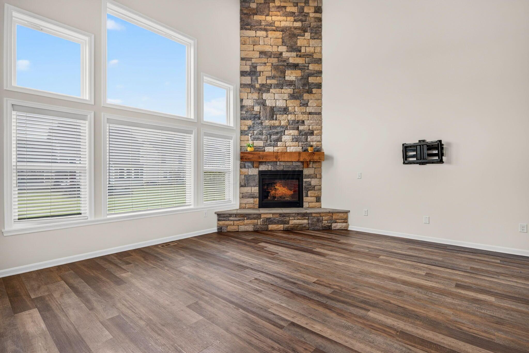 Living room with a tall stone fireplace, large windows, and brown wood-look flooring.