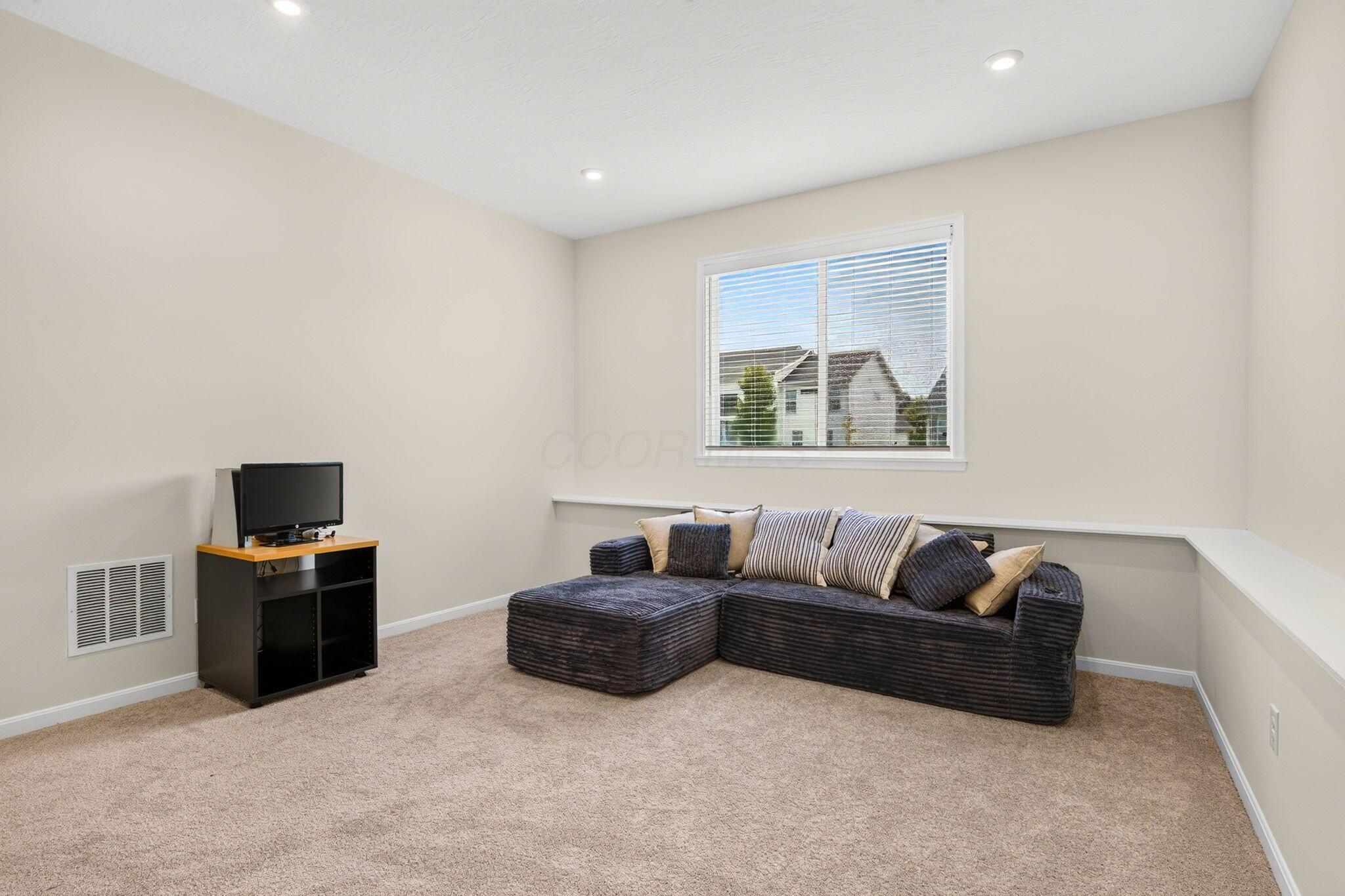Carpeted living area with a dark sectional sofa, television, and window with blinds.