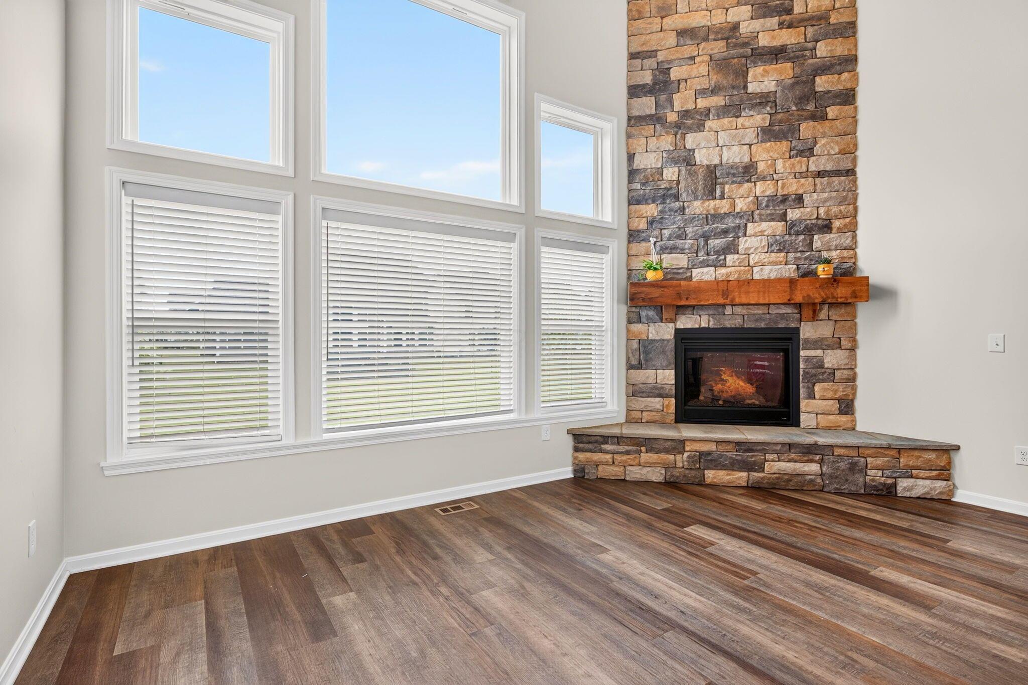 Vacant living room with a tall stone fireplace, large windows, and dark wood flooring.