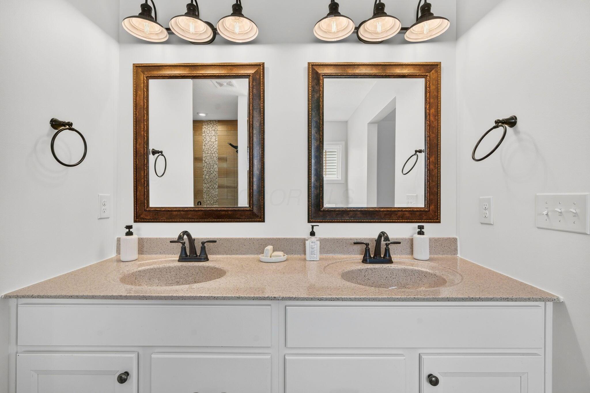 Bathroom double vanity with speckled countertop, bronze faucets, and two ornate framed mirrors.