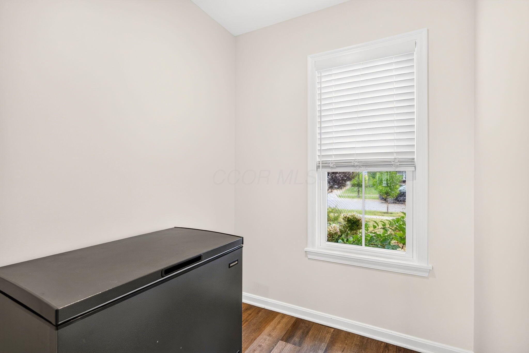 Empty room with wood-look flooring, a window with blinds, and a large chest freezer.