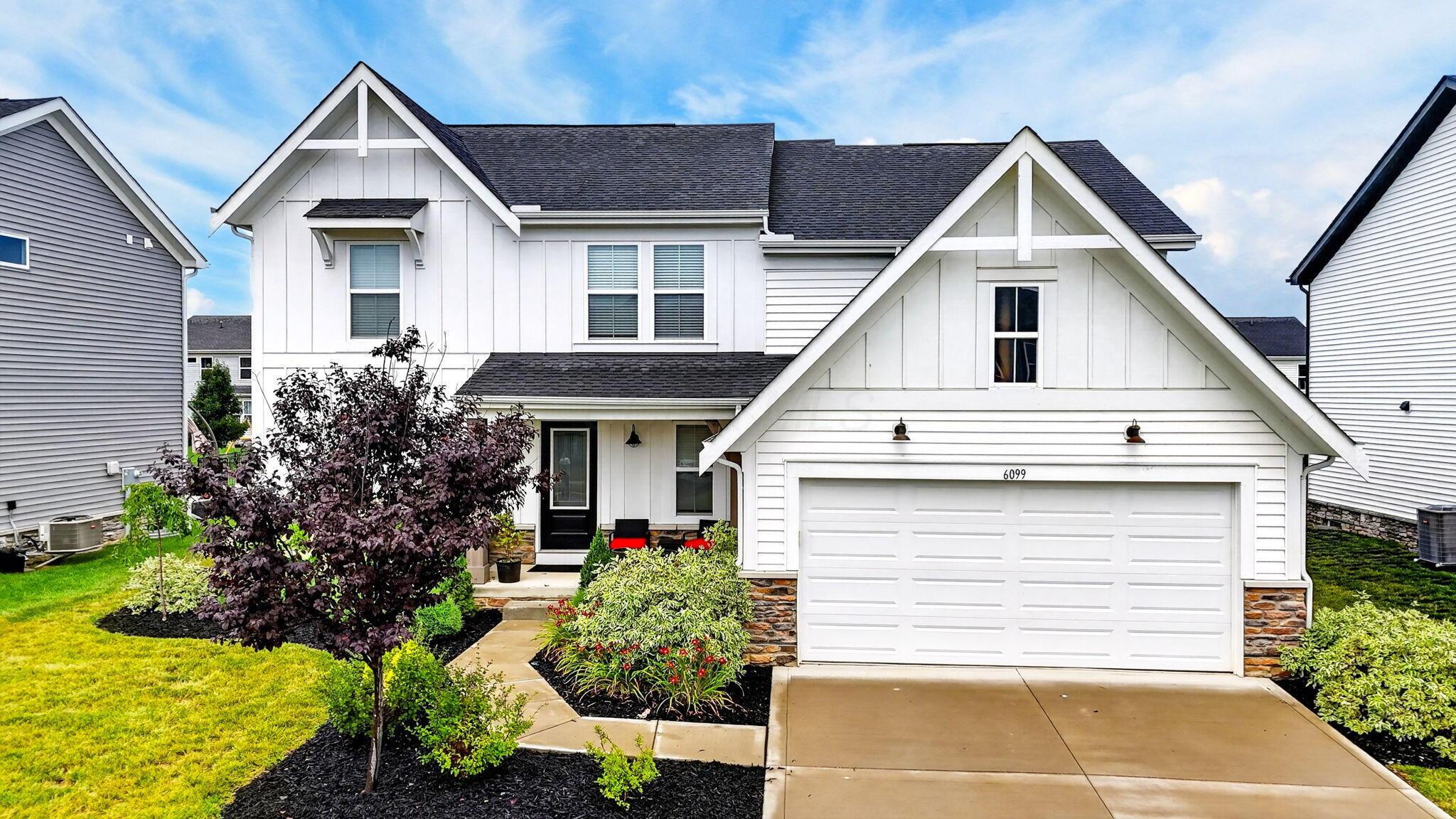 White two-story home with prominent gables, stone accents, landscaped garden beds, and a two-car garage.