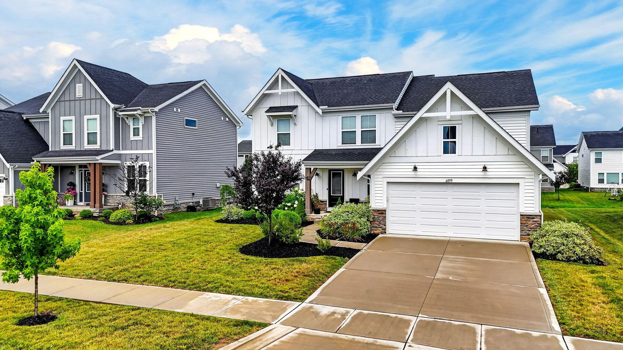 White two-story home with a prominent gabled garage, landscaped front yard, and 6099 above the garage.