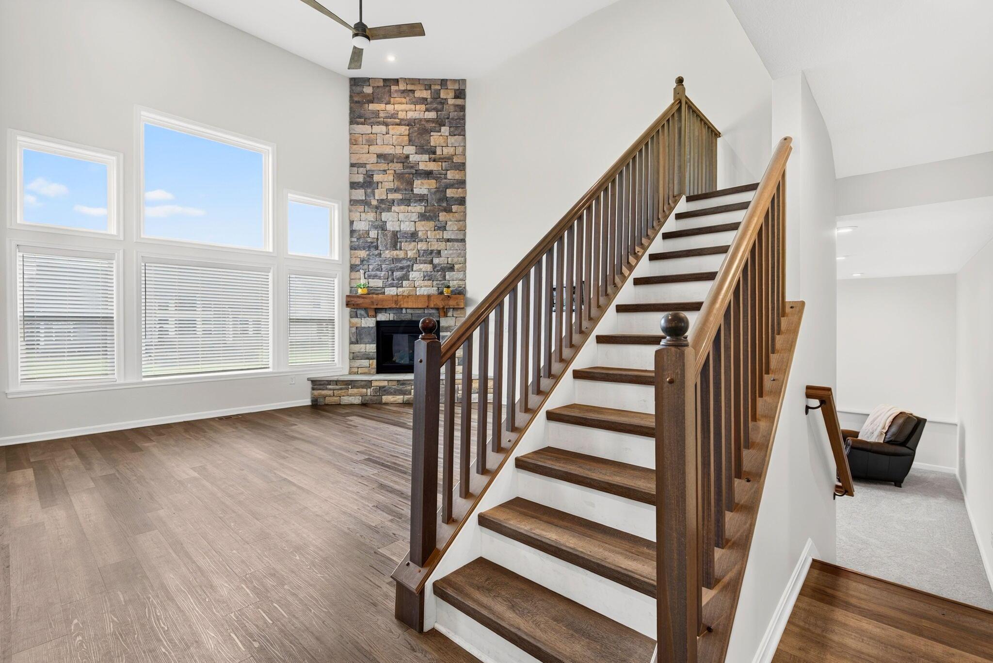 Living room with a tall stone fireplace, large windows, and a dark wood staircase.