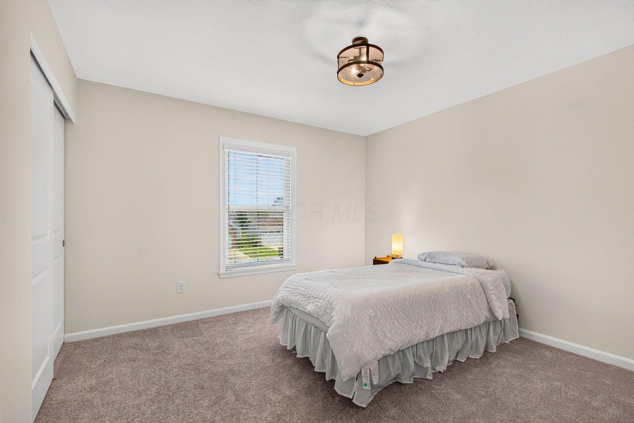 Bedroom with a white bed, taupe carpet, and a window with blinds.