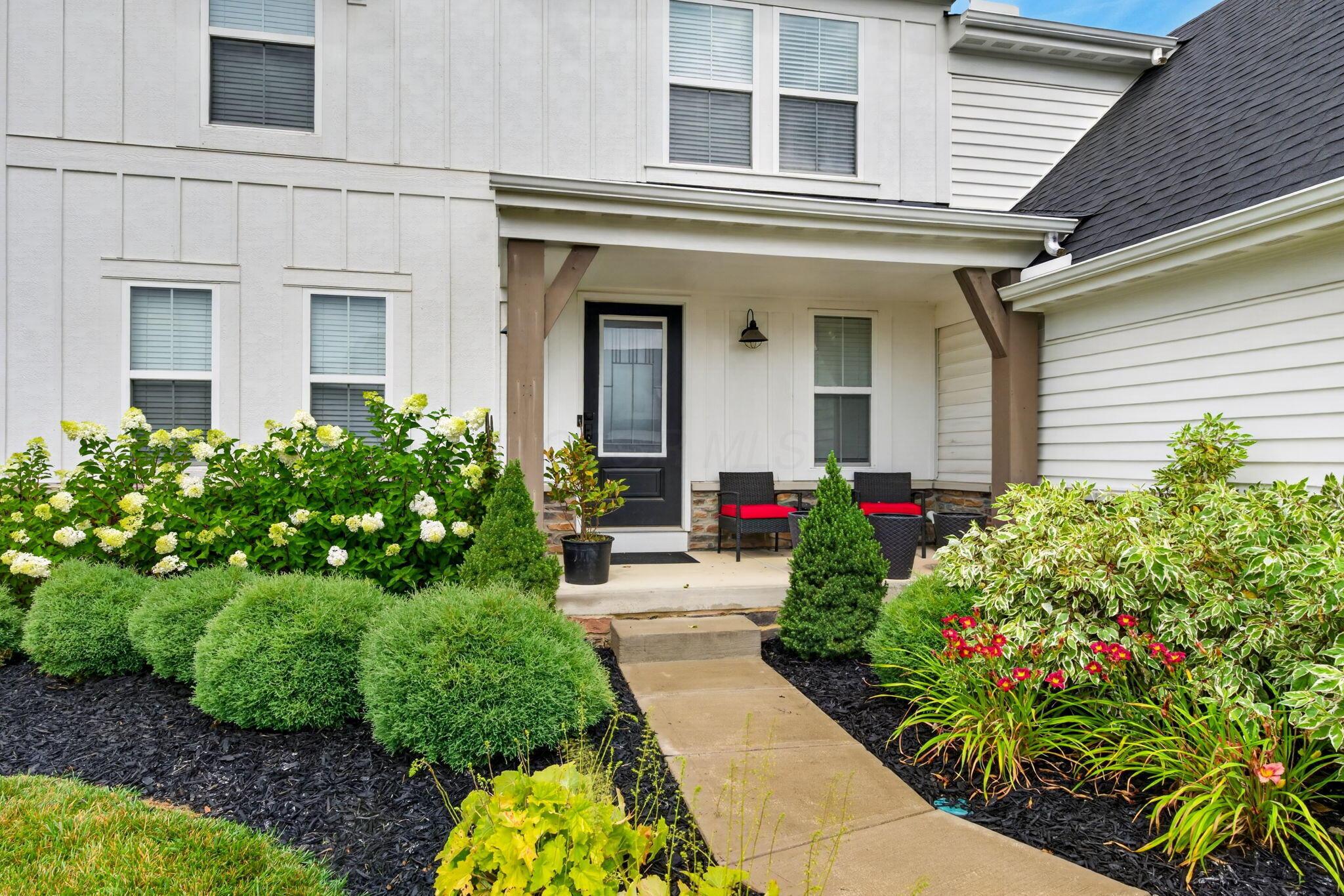 White home’s covered front porch with a black door, red-cushioned chairs, and lush flower beds.