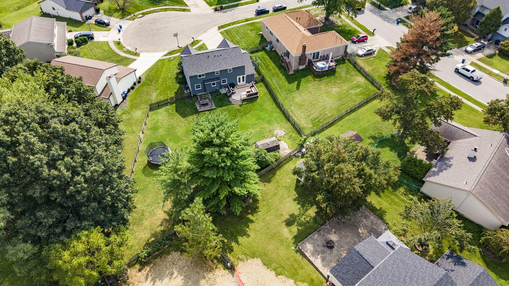 Aerial view of a blue two-story home with a deck, fenced backyard, trampoline, and mature trees.