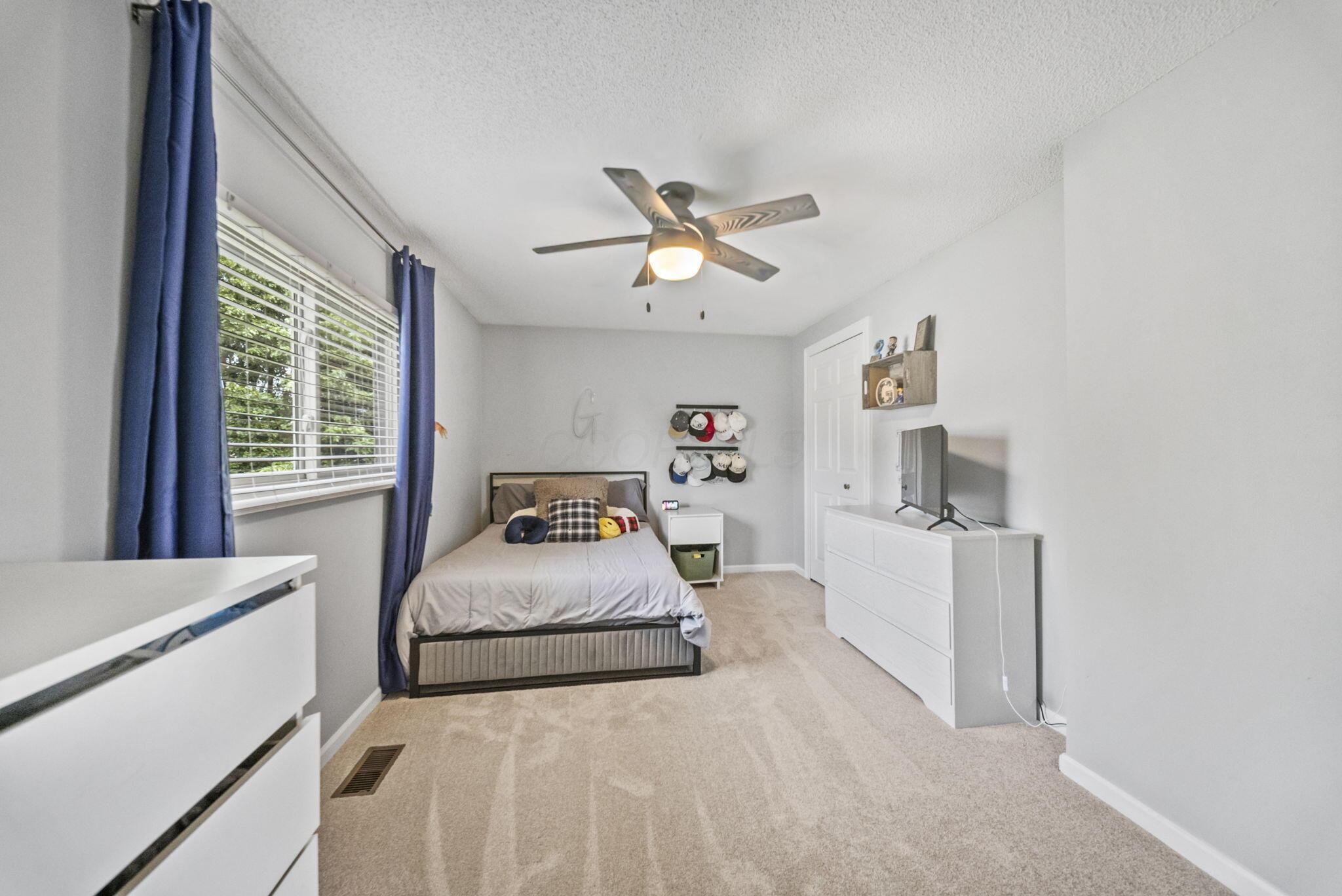 Bedroom with a bed, blue curtains, beige carpet, and a ceiling fan with its light on.