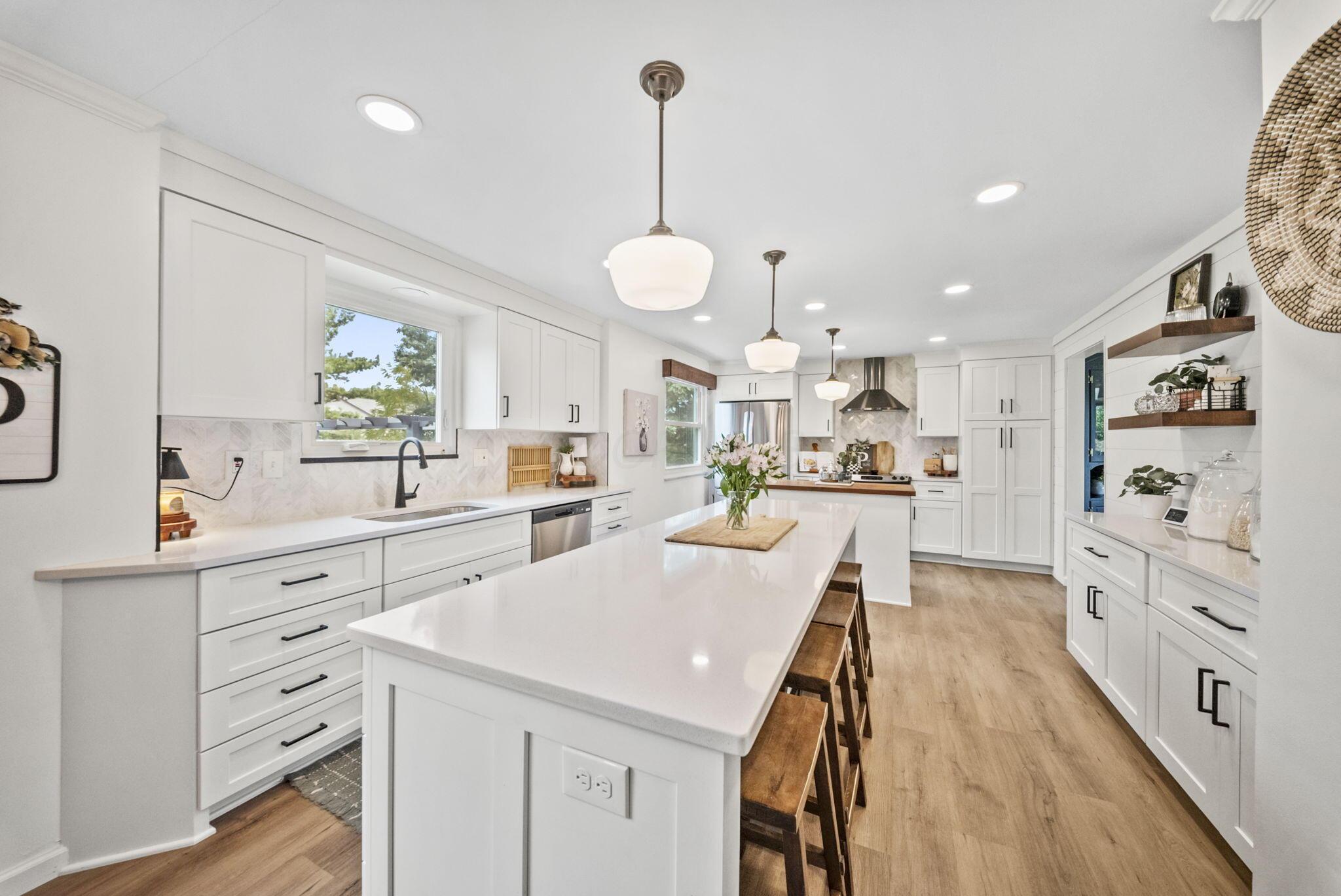 Bright white kitchen with a large island, wooden stools, pendant lights, and warm wood floors.
