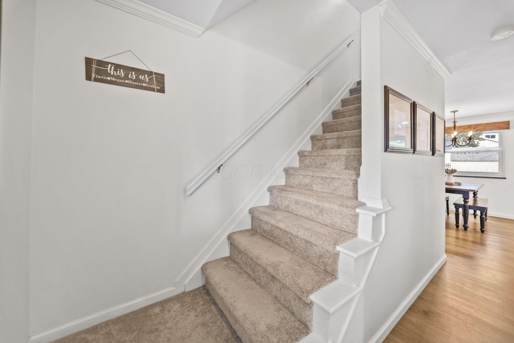 Carpeted staircase with white handrail; framed pictures and a dining area are visible beside it.