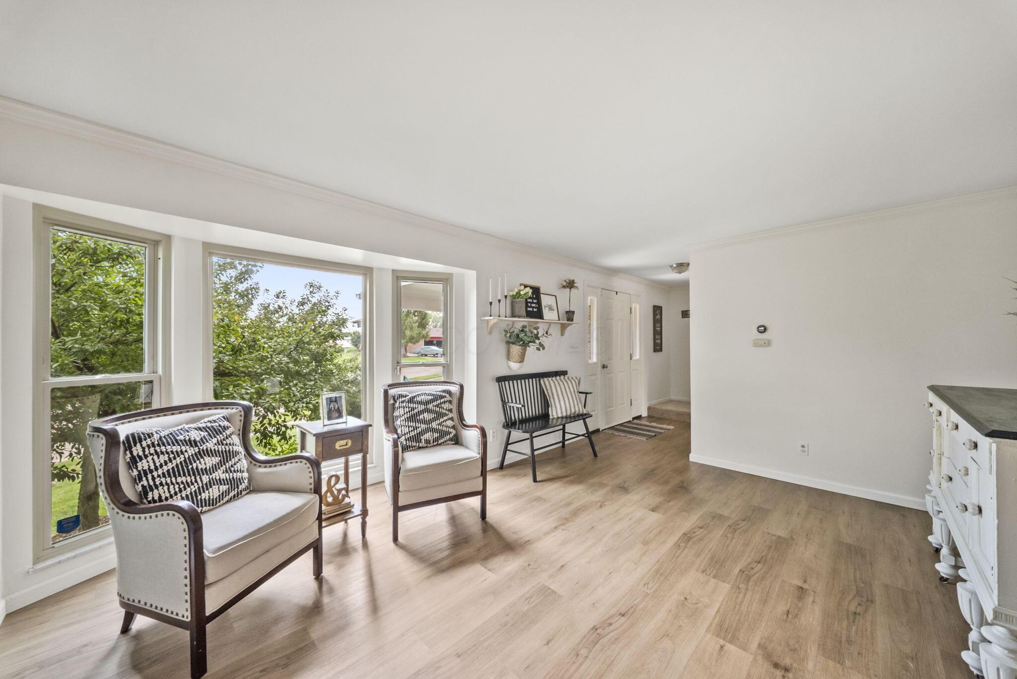 Staged living room with large leafy-view windows, upholstered chairs, black bench, and light wood floors.