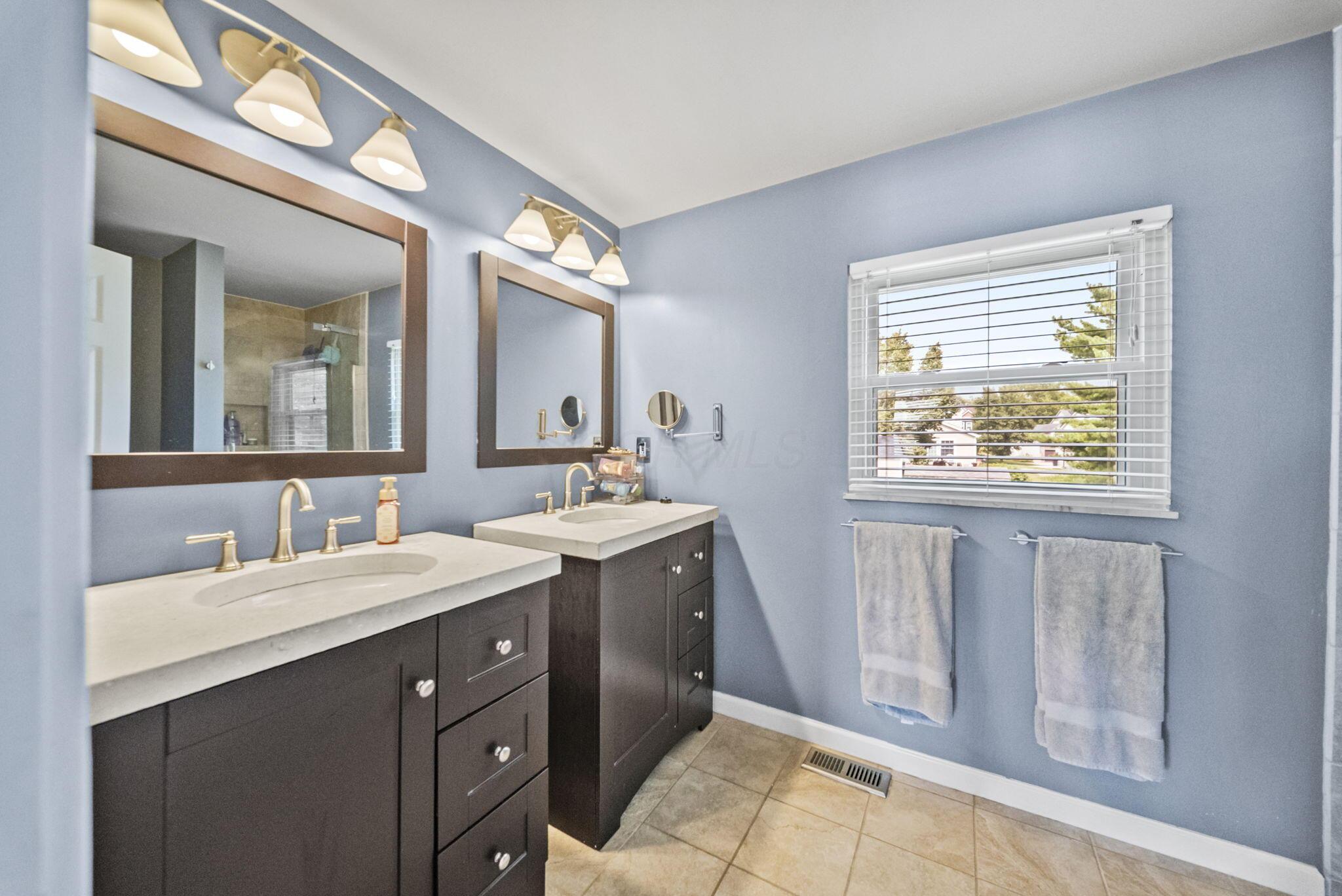 Bathroom with dual vanities, framed mirrors, pale blue walls, and a window with white blinds.