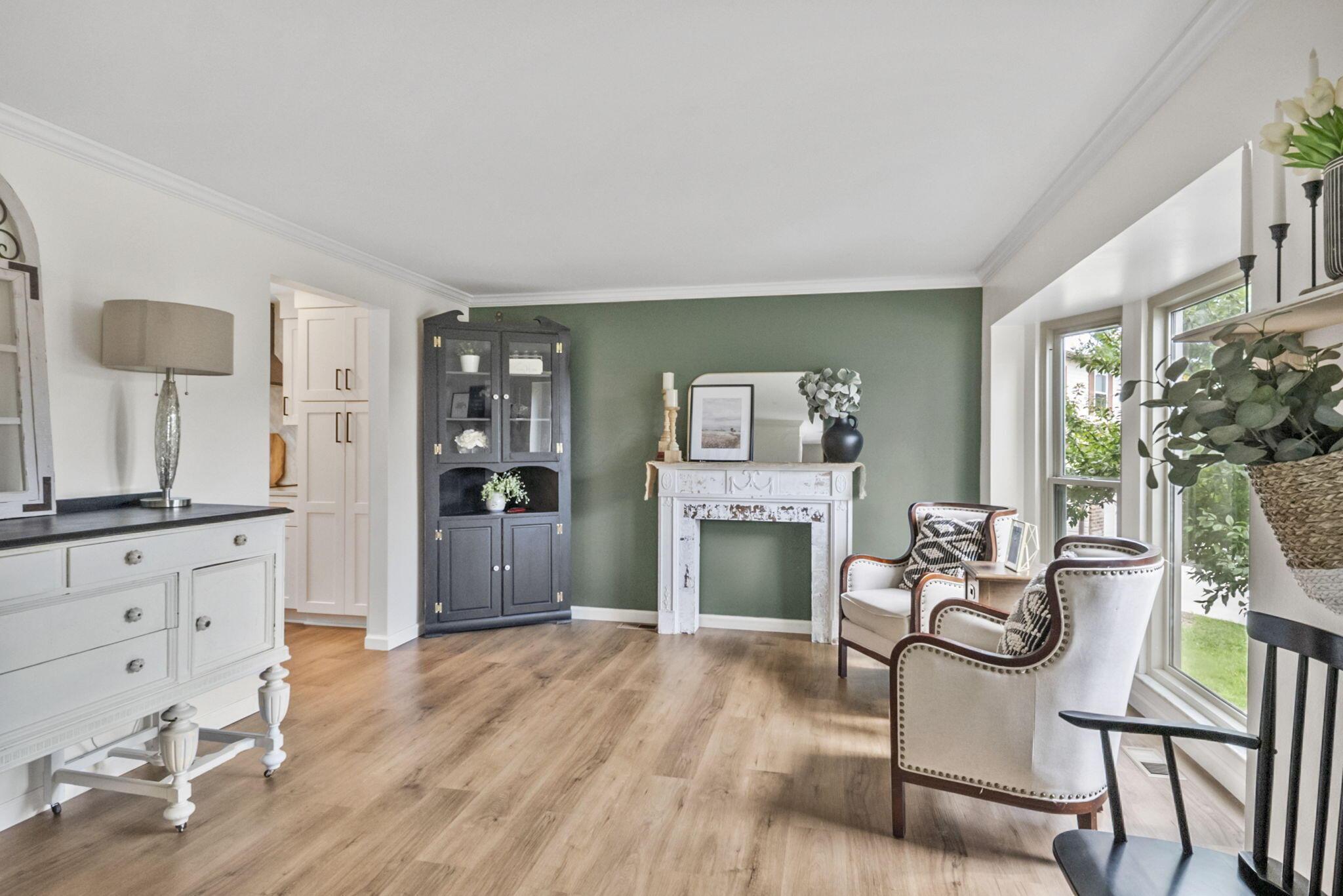 Living room with a sage-green wall, white mantel, wood floors, and upholstered chairs beside tall windows.