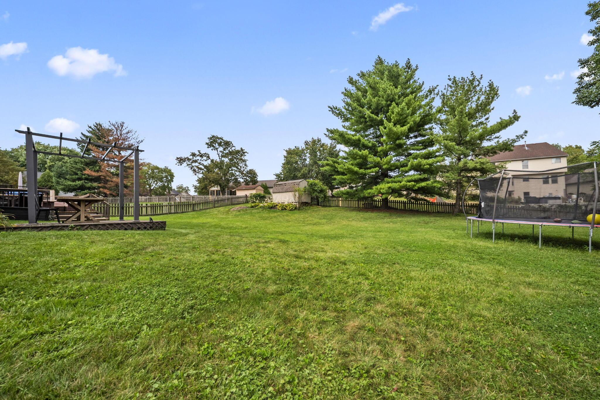 Spacious fenced backyard with a broad lawn, deck and pergola, mature trees, and a trampoline.