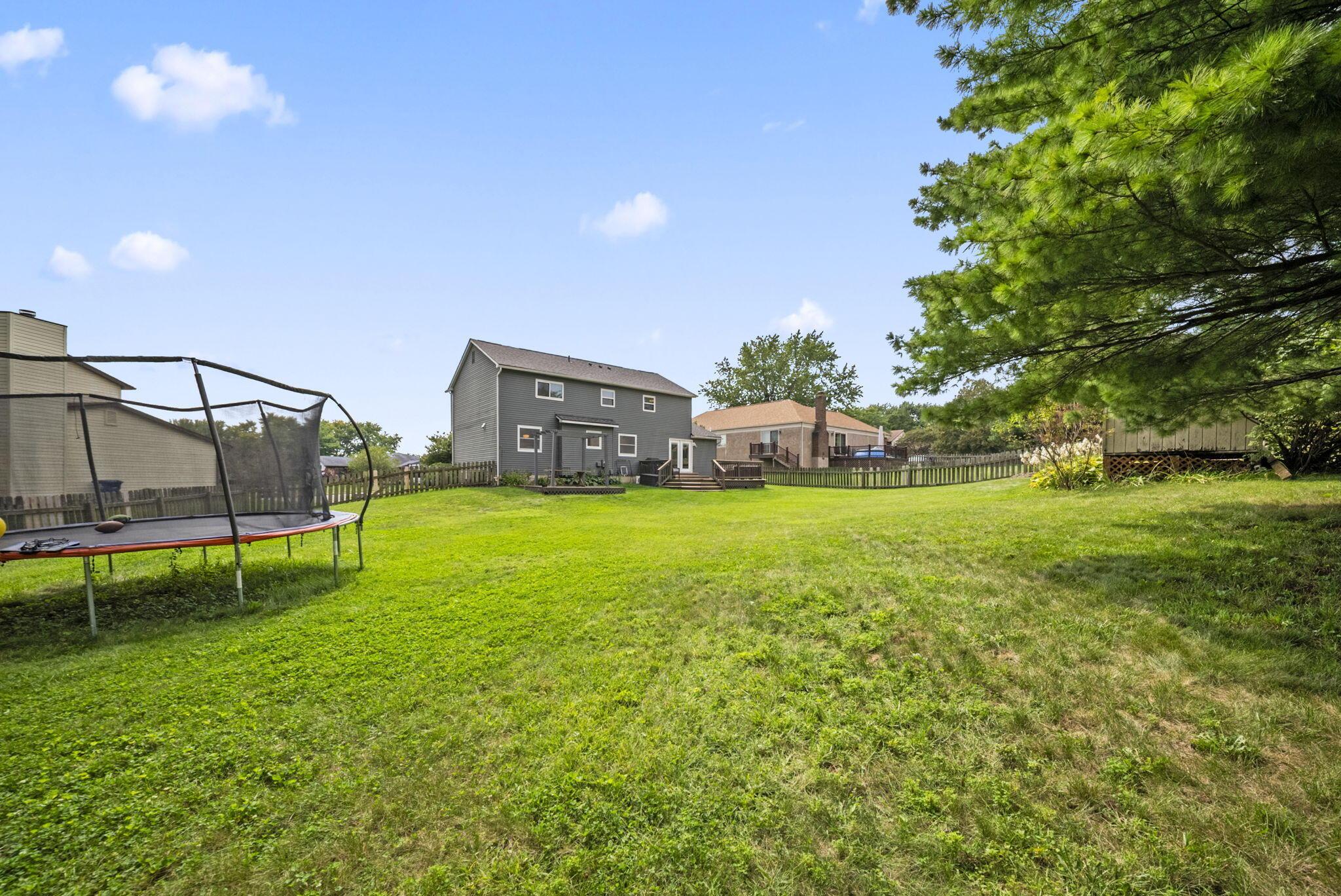Spacious fenced backyard with a trampoline, gray two-story home, and large pine tree.