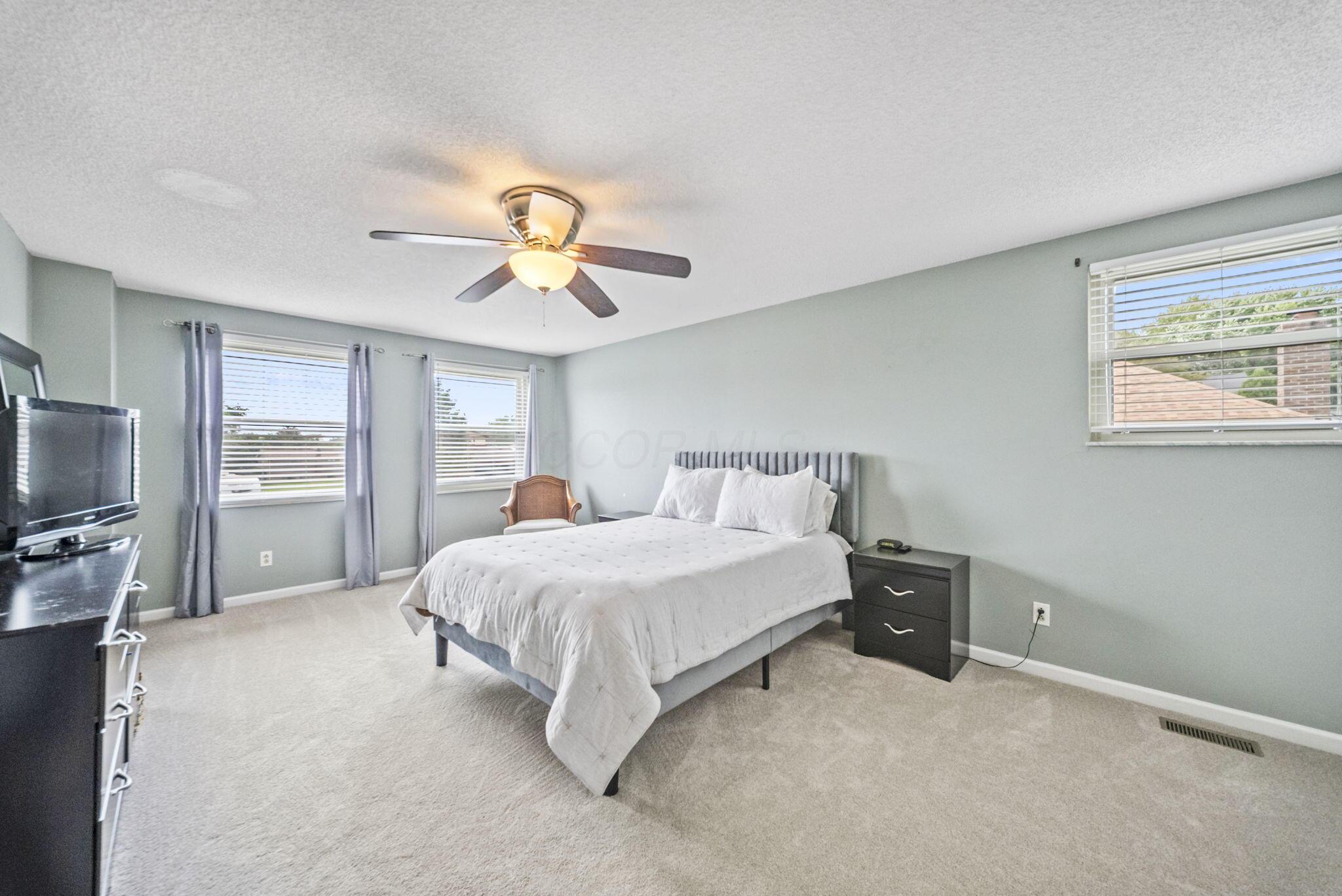 Furnished bedroom with a white bed, gray headboard, ceiling fan, and three windows.