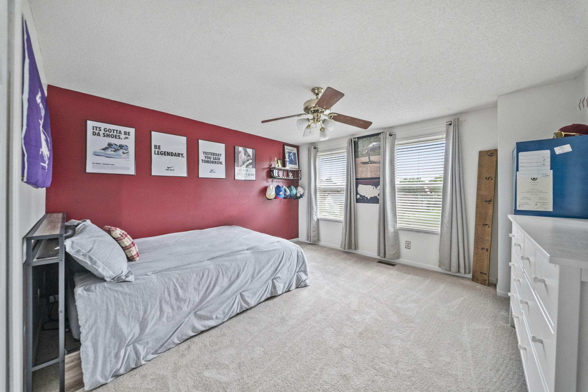 Bedroom with a red accent wall, sneaker posters, two curtained windows, and a ceiling fan.