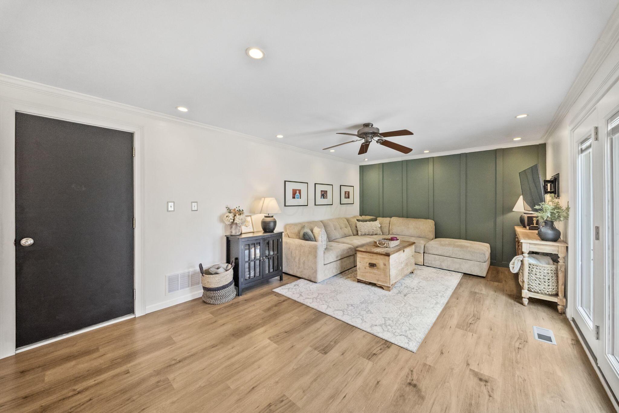 Living room with a beige sectional, green paneled accent wall, light wood floors, and large windows.