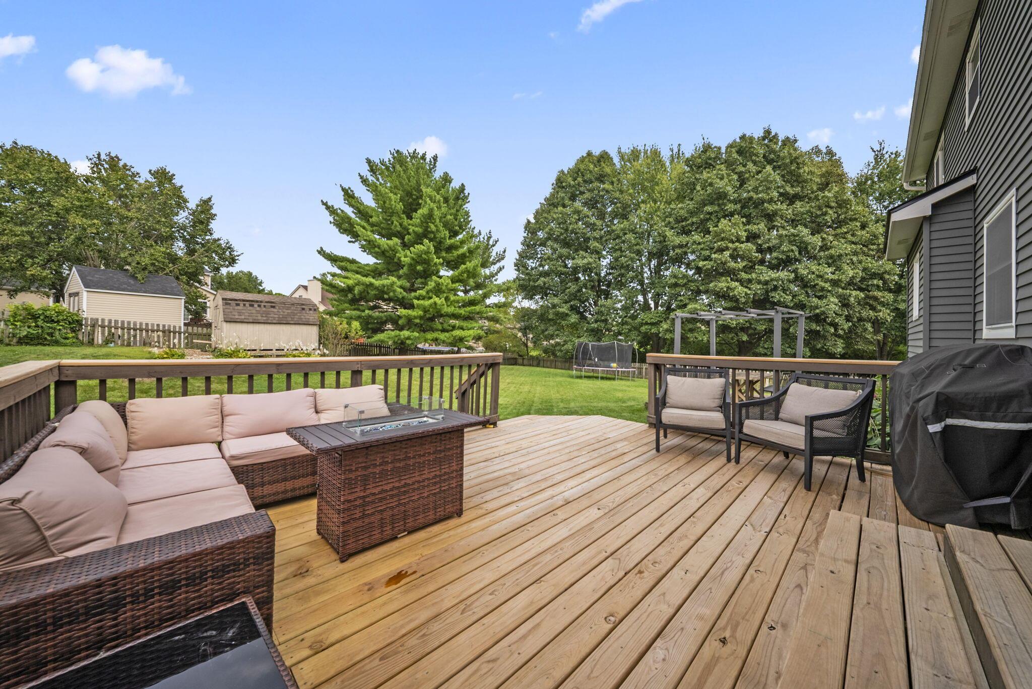 Wooden backyard deck with wicker seating, a fire table, and views of a tree-lined lawn.