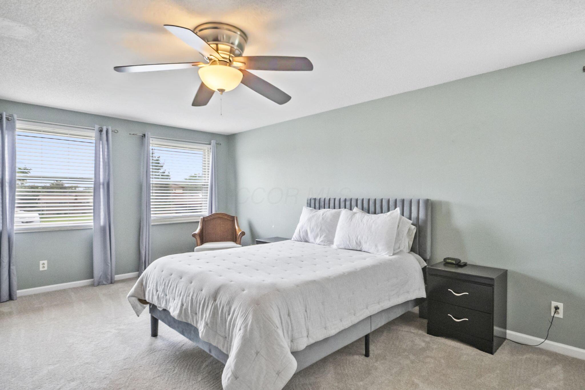 Furnished bedroom with a white bed, gray upholstered headboard, and two curtained windows.