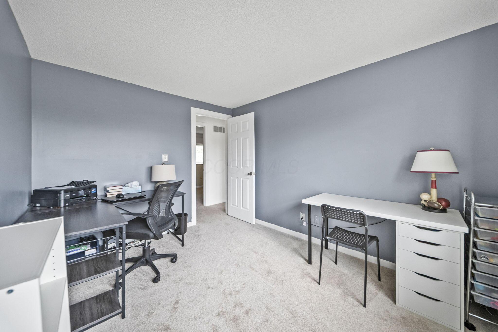 Furnished home office with two desks, gray walls, and an open doorway.
