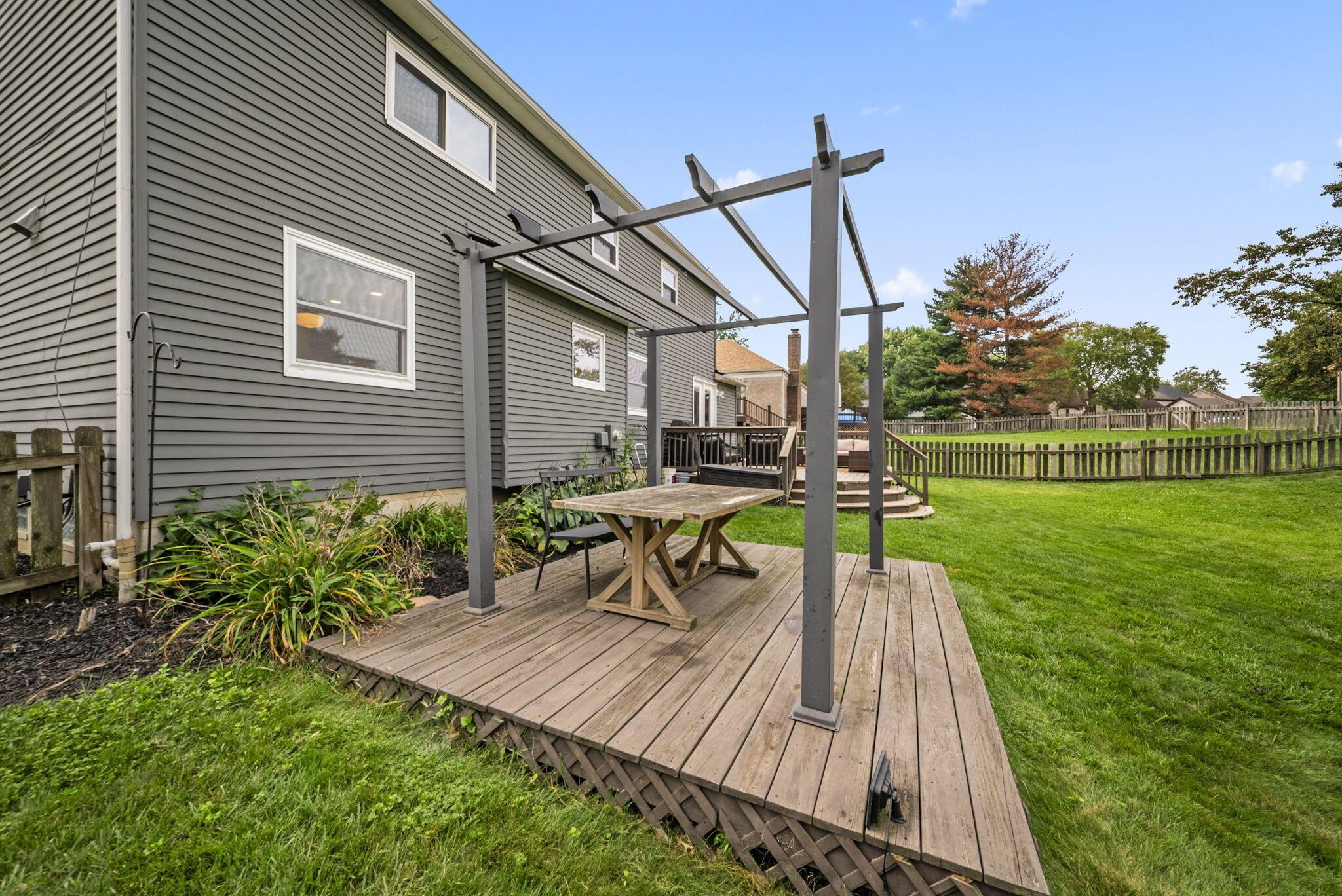 Raised wooden deck with a pergola and outdoor table beside a gray-sided home and fenced lawn.