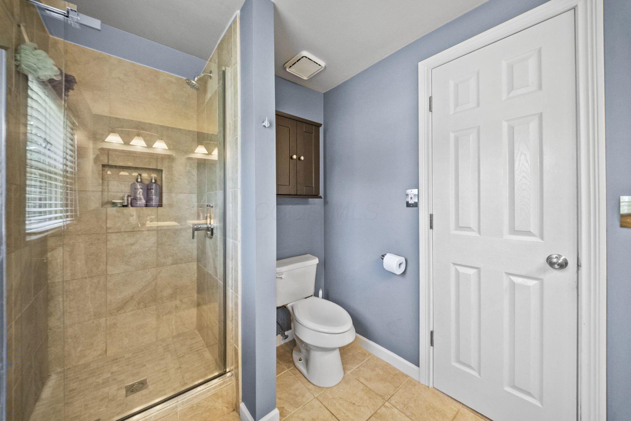Bathroom with a glass-enclosed beige-tile shower, recessed shelf, toilet, and white paneled door.