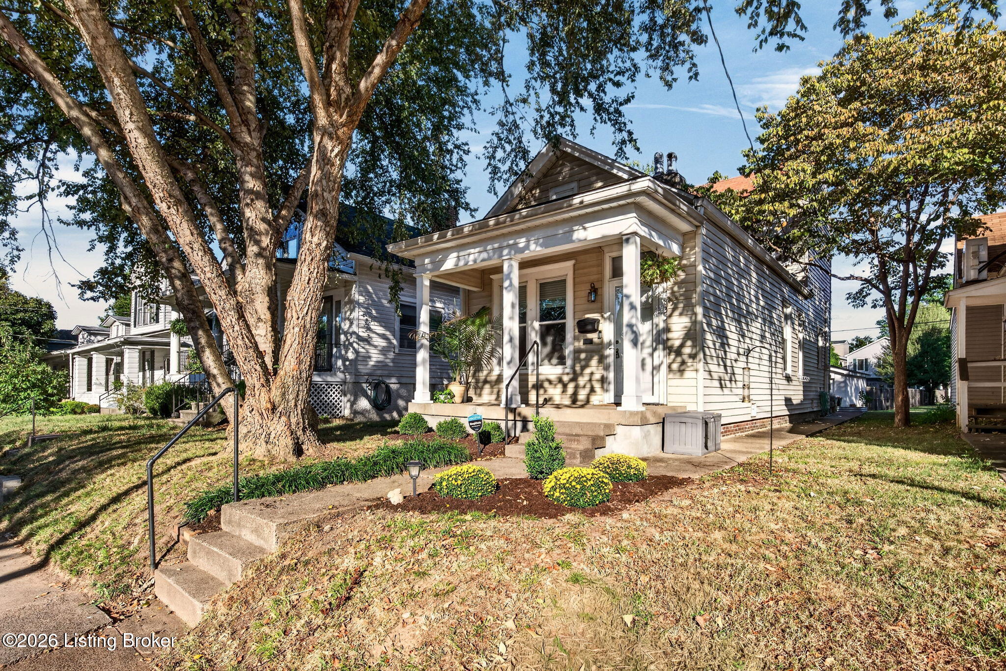 Small bungalow with a covered porch, concrete steps, mature trees, and yellow flowers in the front yard.
