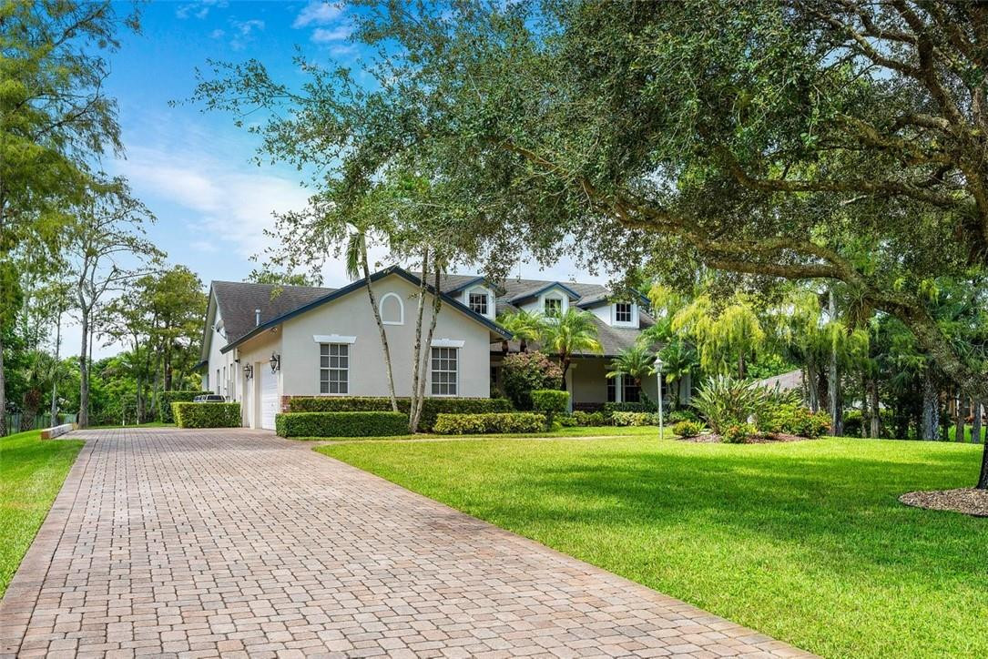 Large light-colored home with a curving brick-paver driveway, mature trees, and tropical landscaping.