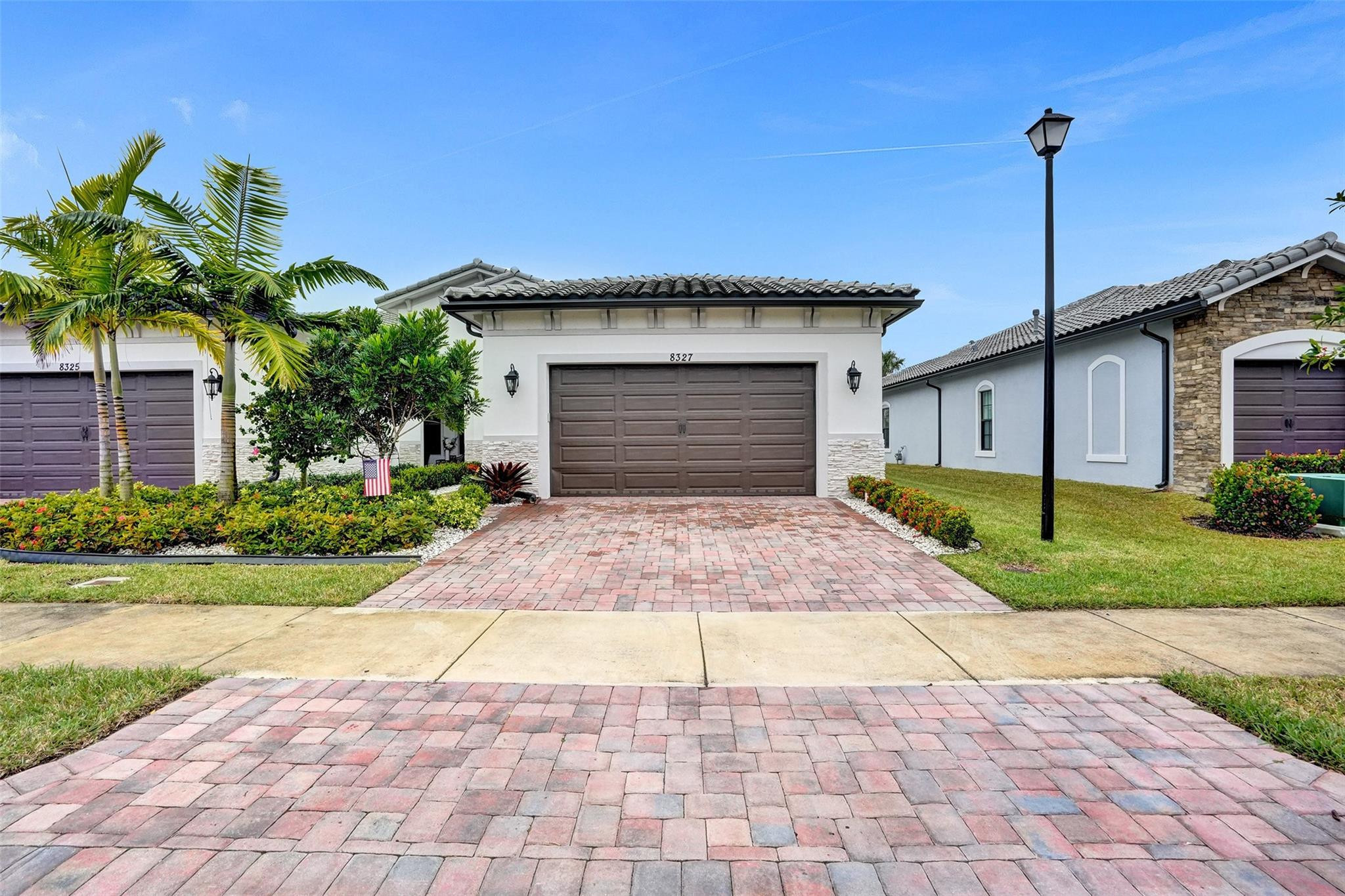 White stucco home with a brown garage door, landscaped yard, and wide brick-paver driveway.