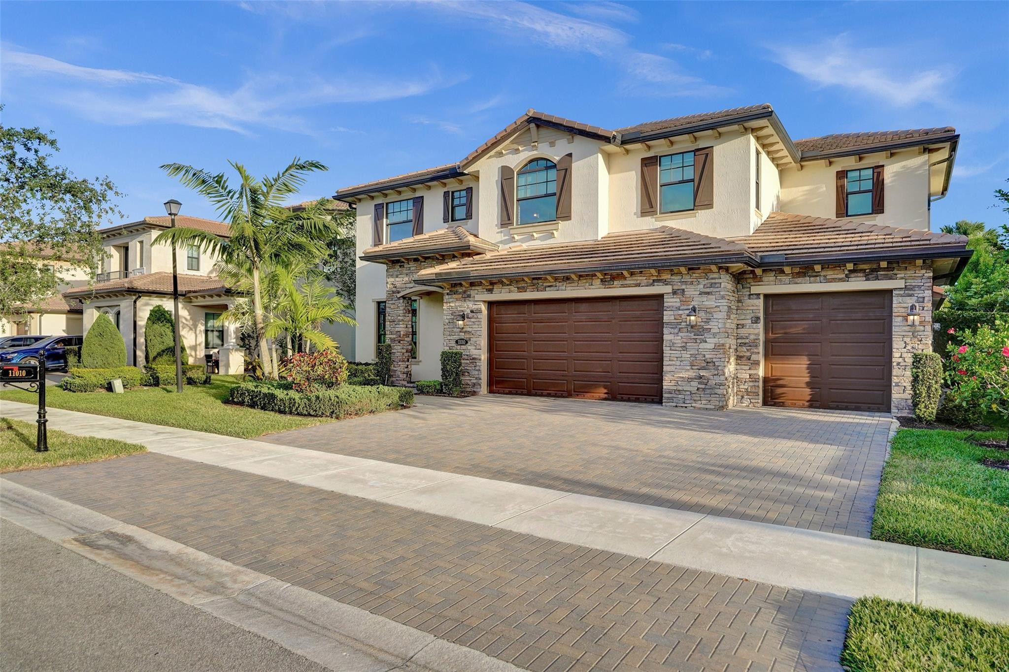 Two-story cream stucco home with stone accents, twin wooden garage doors, and a broad paver driveway.