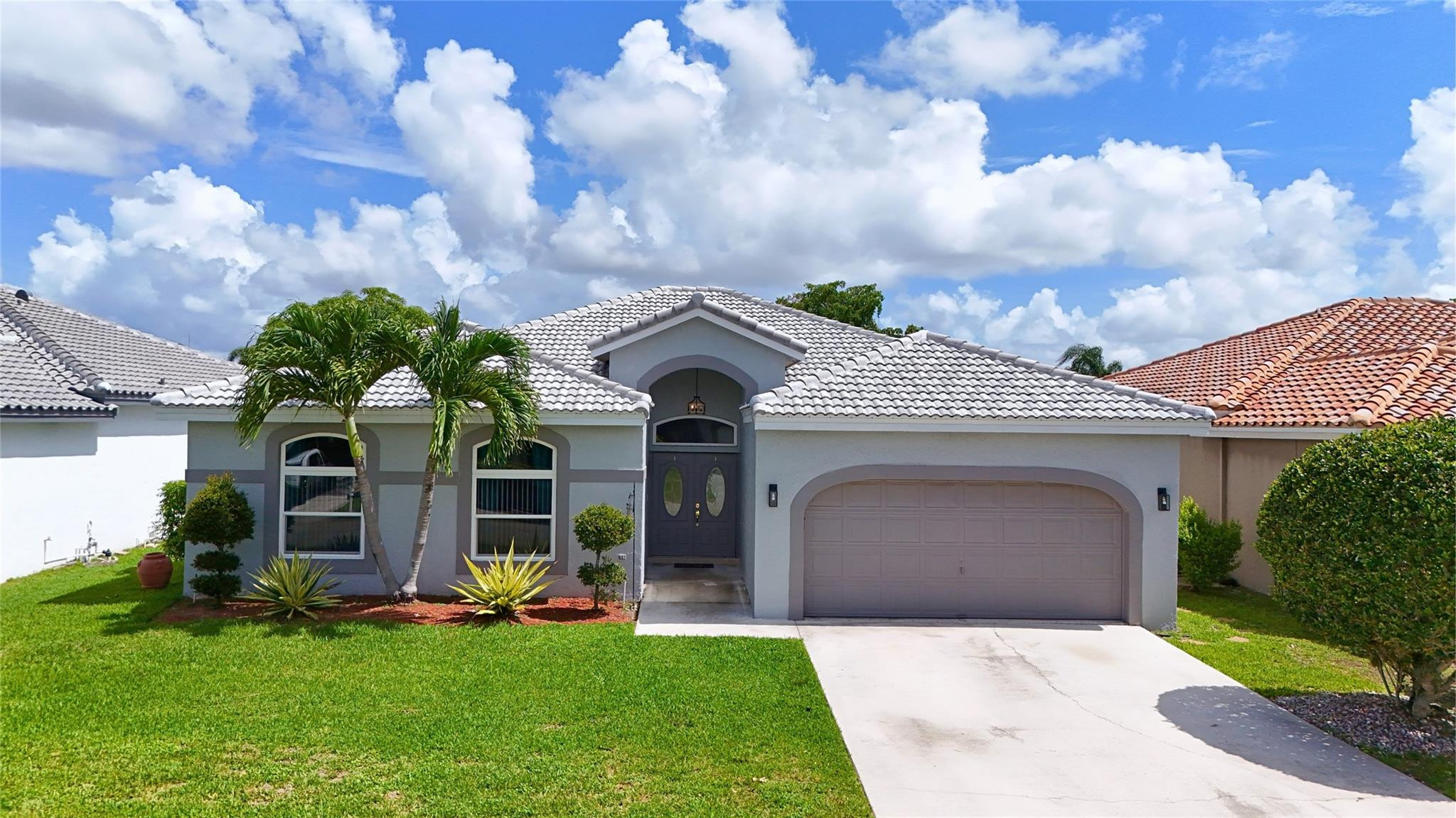 Gray single-story home with a tile roof, arched entry, two-car garage, and palm trees in the front yard.
