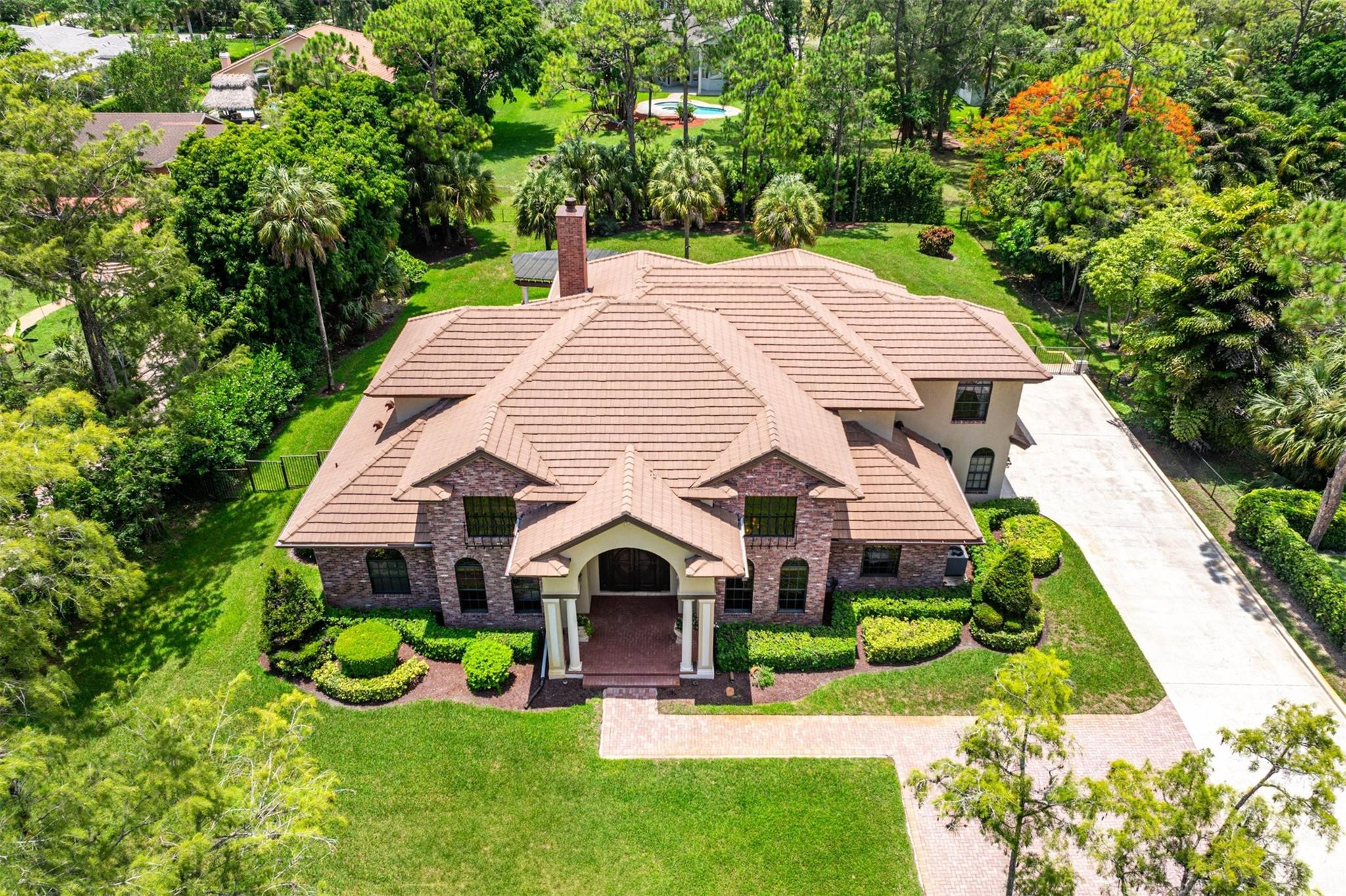 Aerial view of a brick-and-stucco home with a broad tan tiled roof, landscaped lawn, and long driveway.