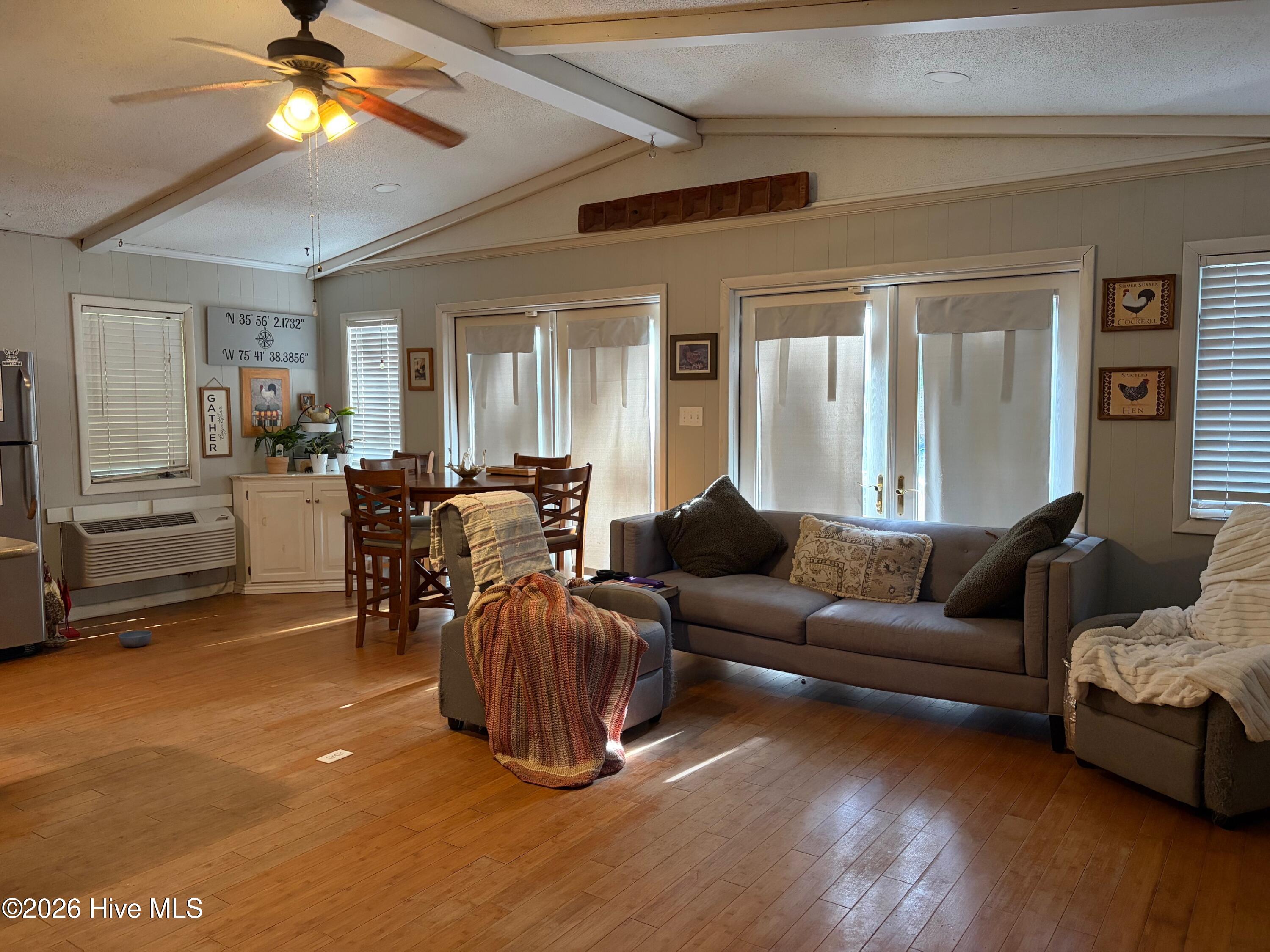 Living and dining room with hardwood floors, vaulted ceiling, gray sofa, and curtained sliding glass doors.