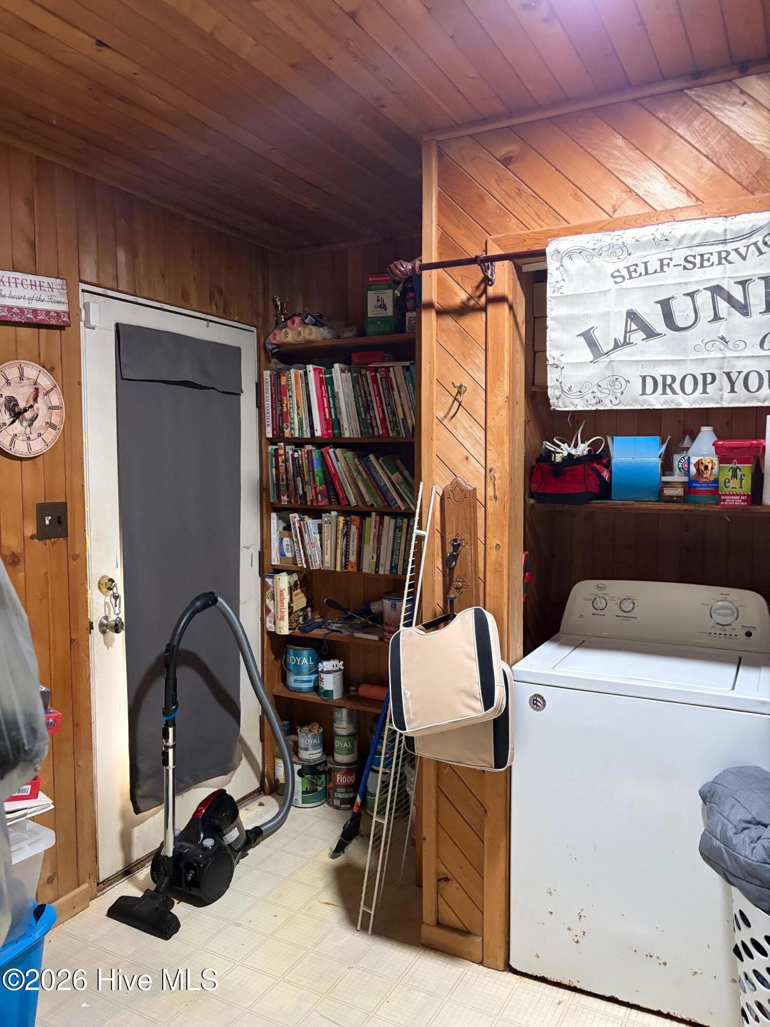 Wood-paneled laundry room with a white washing machine, vacuum, and shelves of books and supplies.