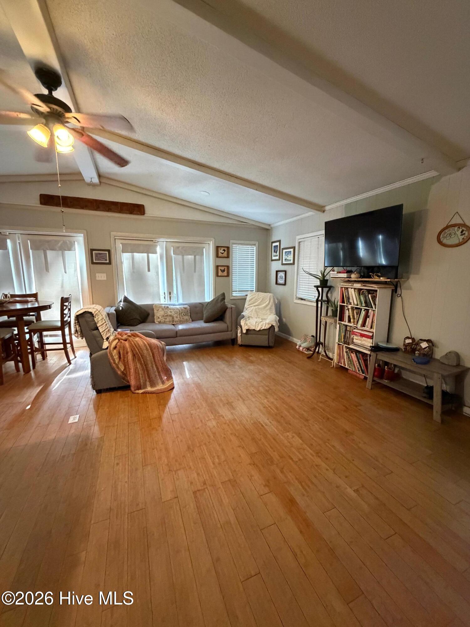 Living room with vaulted beamed ceiling, wood floors, gray seating, and a TV above a bookcase.