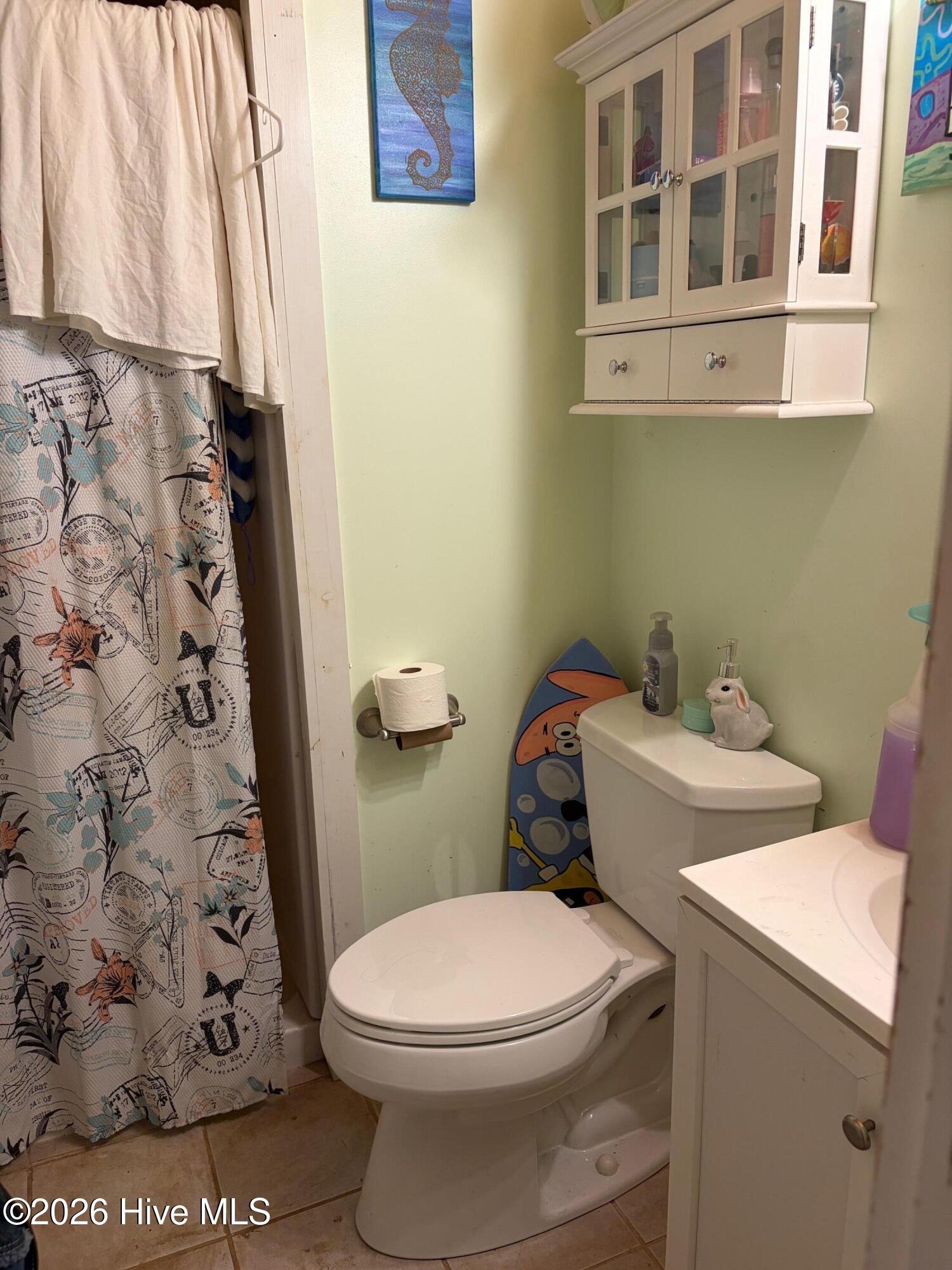 Bathroom with a glass-front wall cabinet, patterned shower curtain, and colorful surfboard beside the toilet.