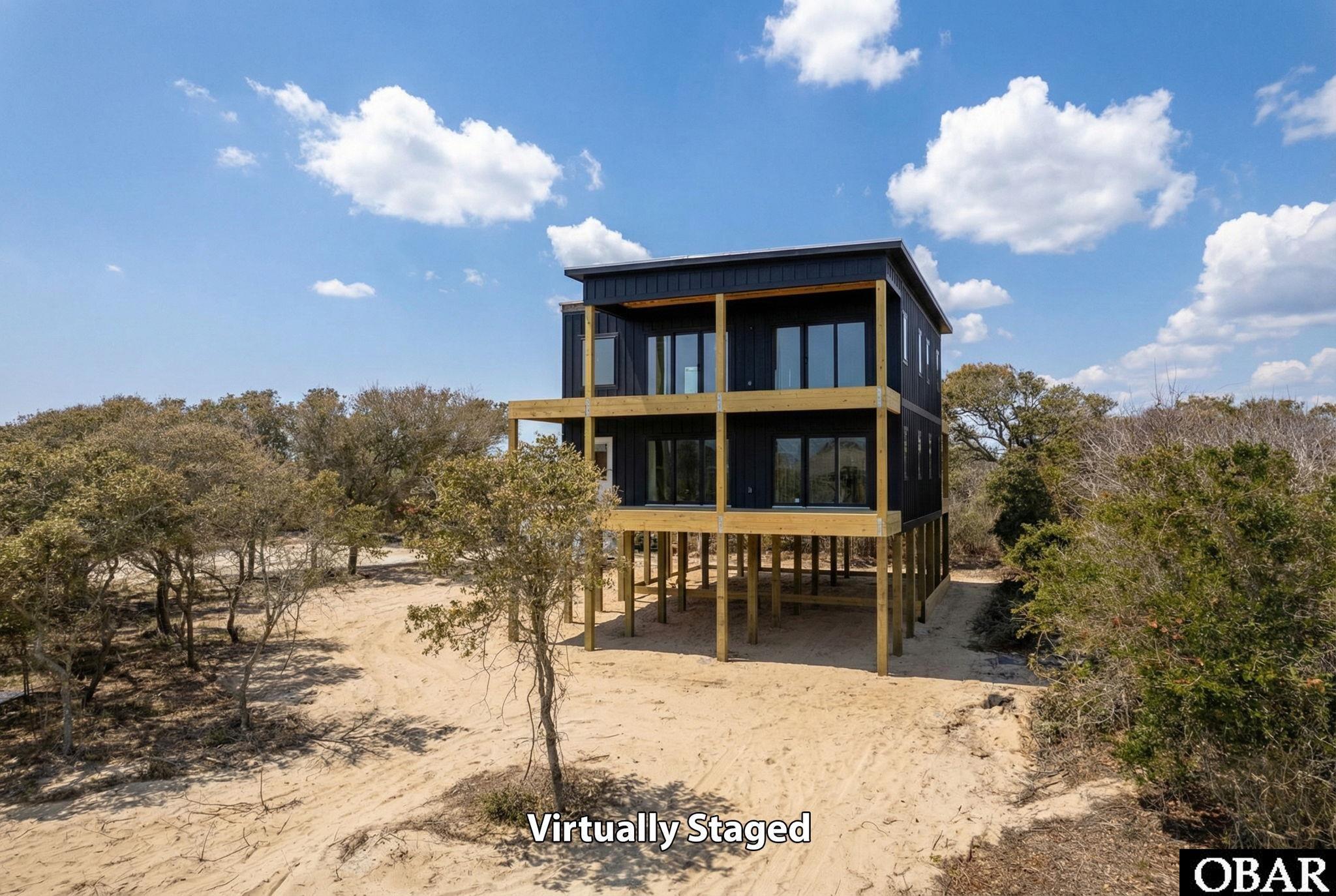 Virtually staged modern coastal home raised on pilings, with wood-framed decks amid sandy ground and coastal trees.