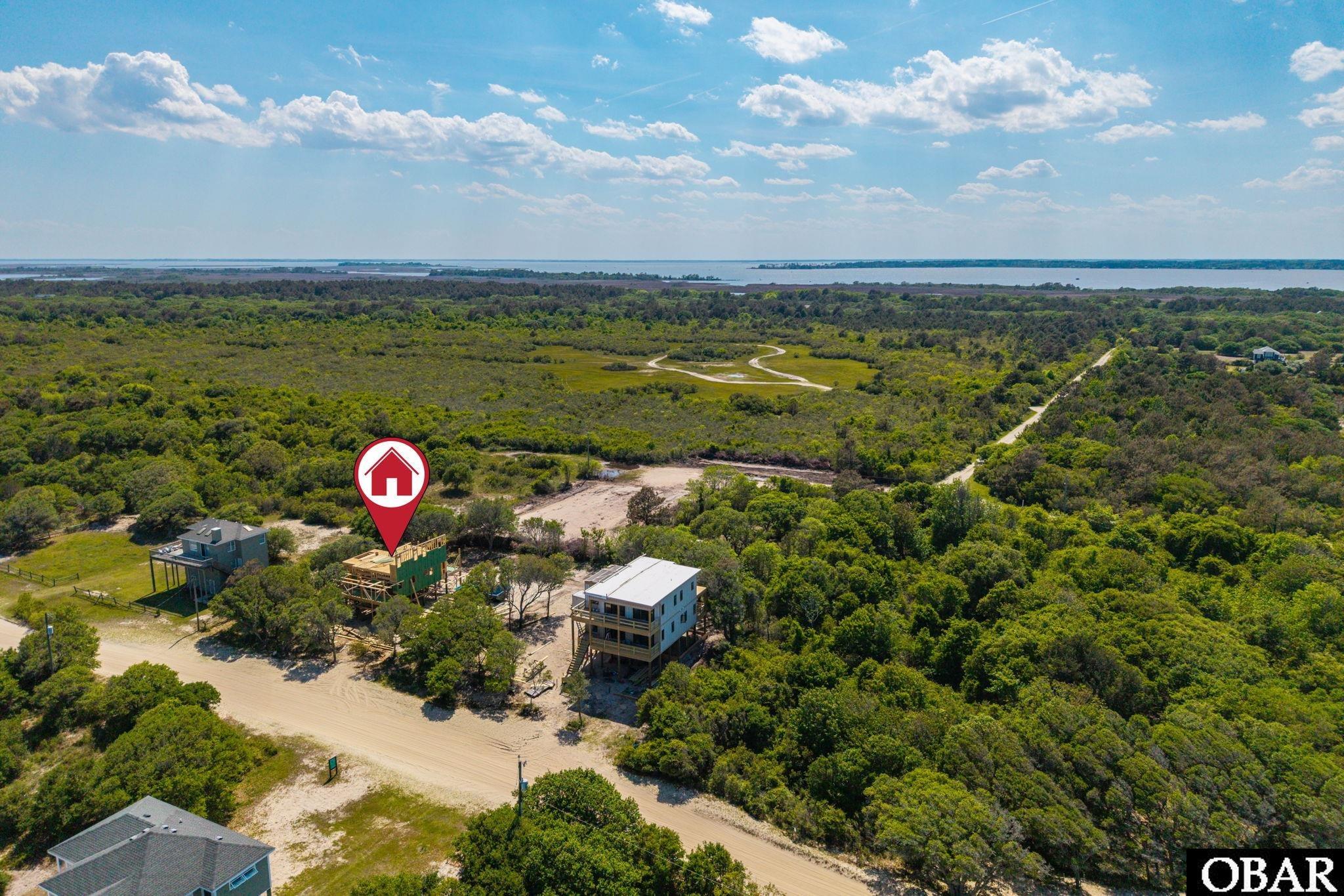 Aerial view of a house under construction on a wooded lot, with water visible on the horizon.