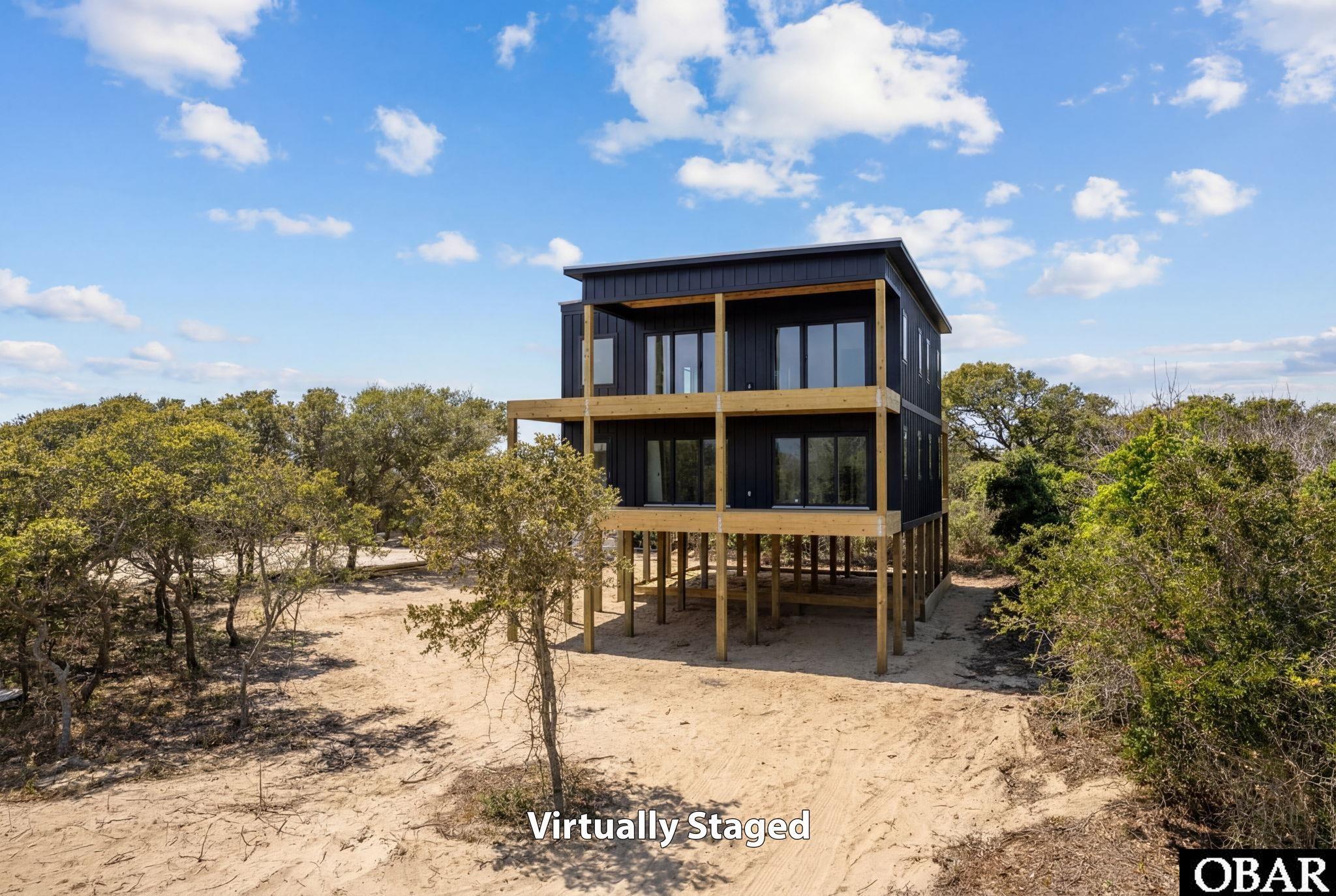 Elevated dark-sided house with wood balconies on a sandy lot surrounded by trees.