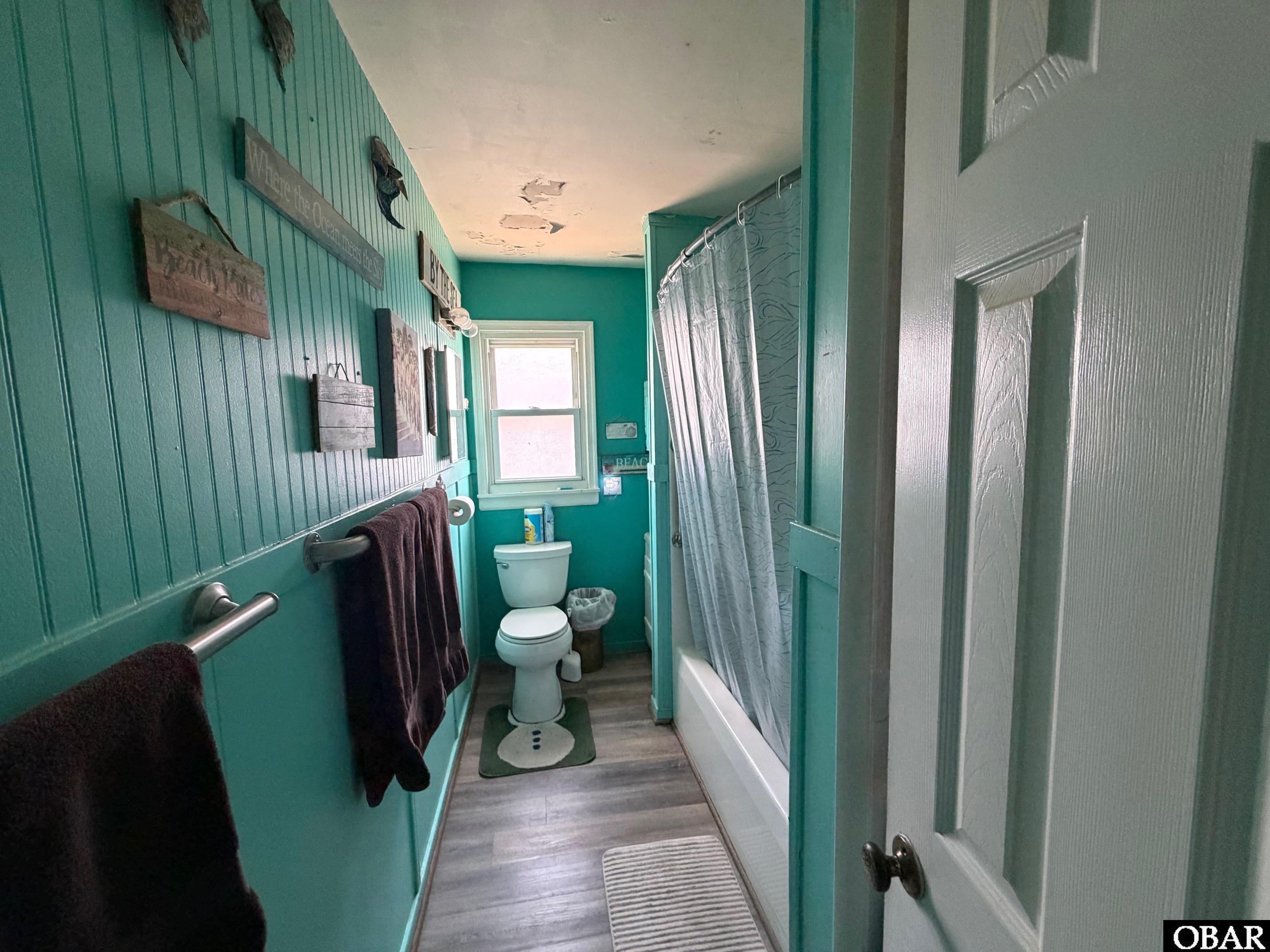 Teal bathroom with a toilet beneath a window, bathtub, patterned shower curtain, and damaged ceiling.