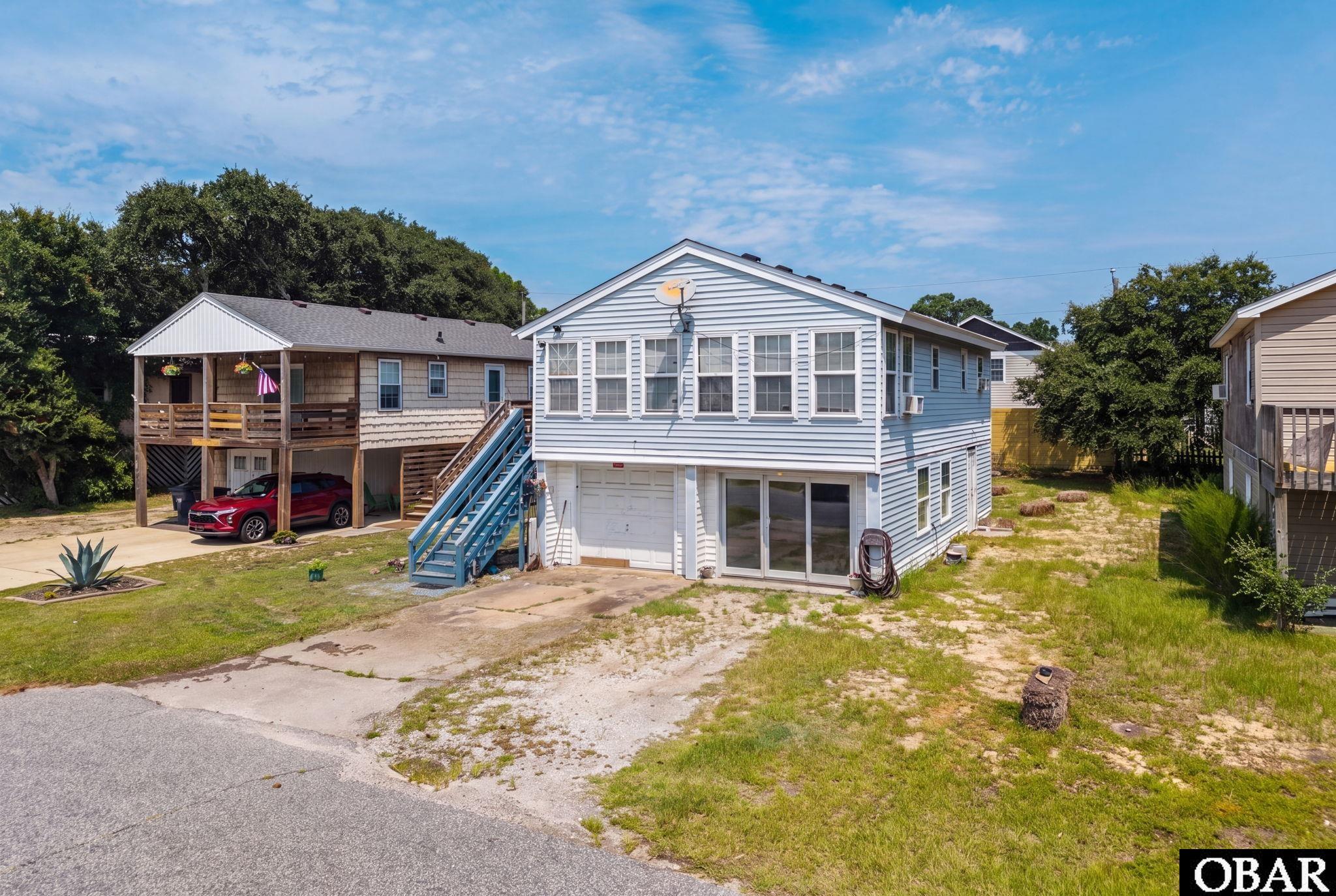 Light blue raised coastal home with exterior stairs, garage, and a sandy grassy lot.
