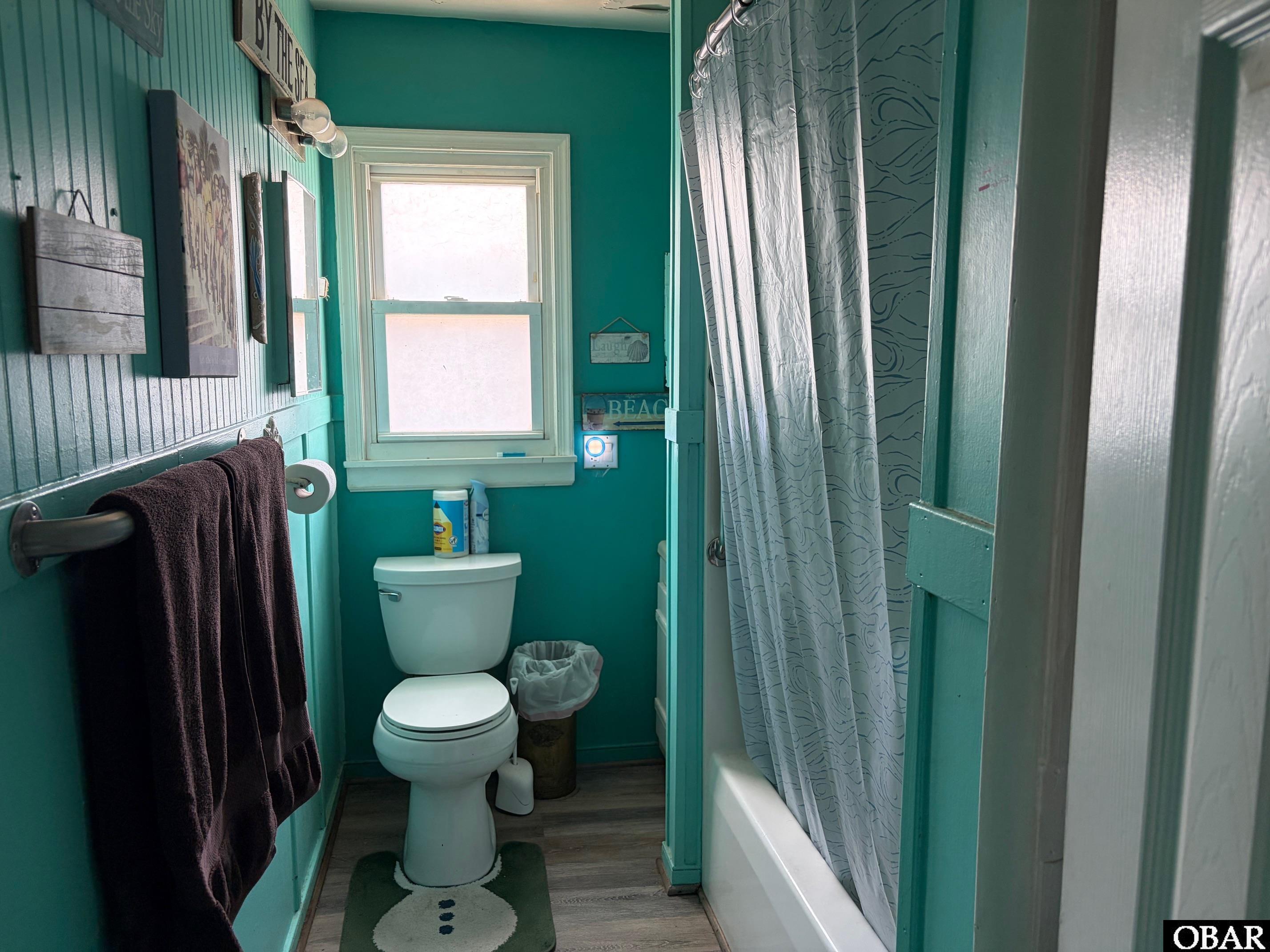 Turquoise bathroom with a toilet beneath a window and a bathtub behind a patterned shower curtain.